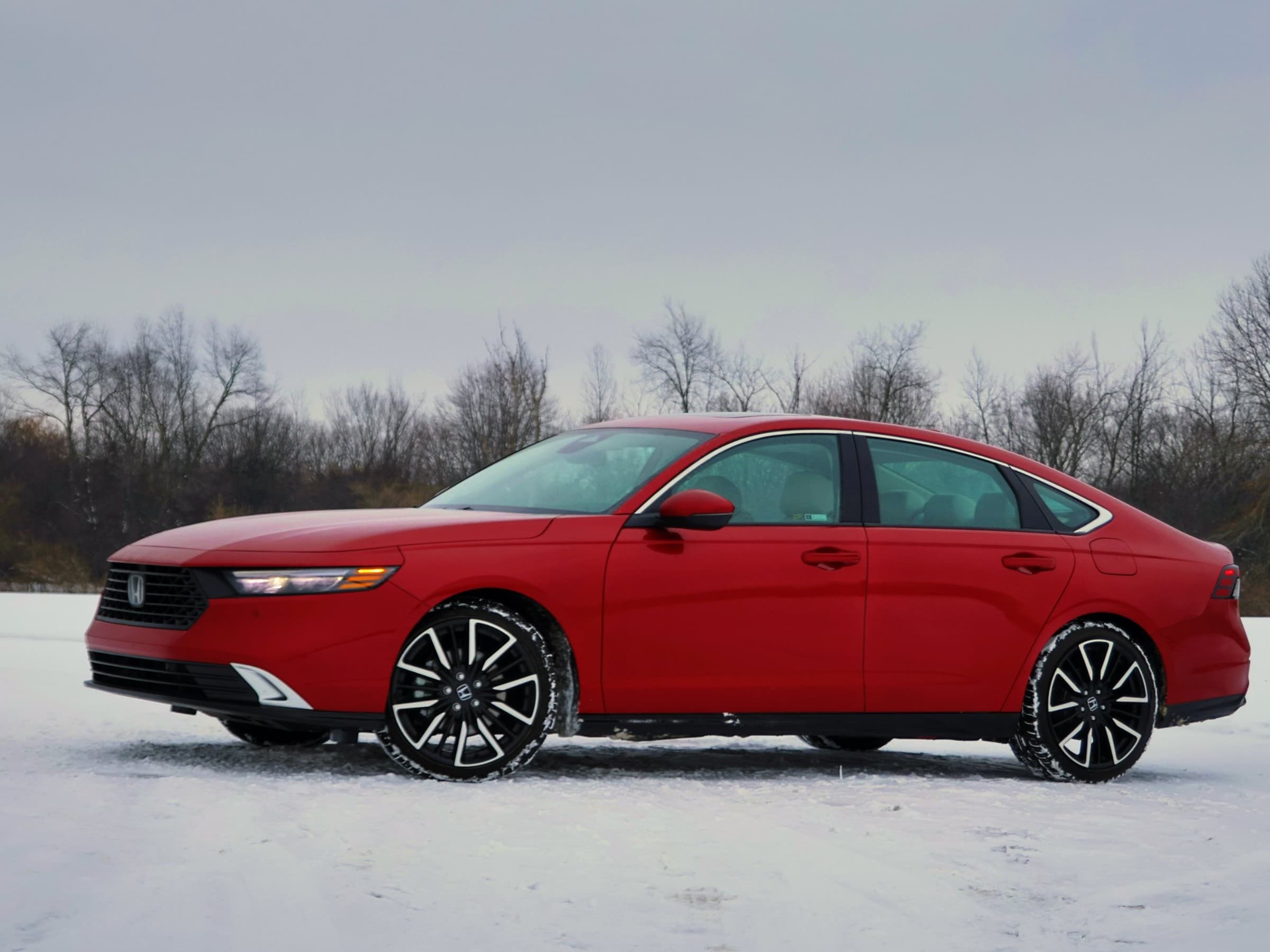 Front three-quarter view of a red Honda Accord Hybrid showing its slim headlights, black grille and long fastback roofline, parked on snow.