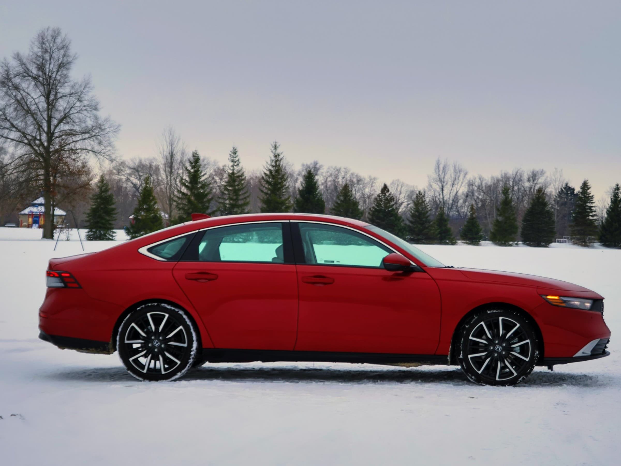 Side profile of a red Honda Accord Hybrid sedan with two-tone wheels, parked on snow in front of a row of evergreen trees.
