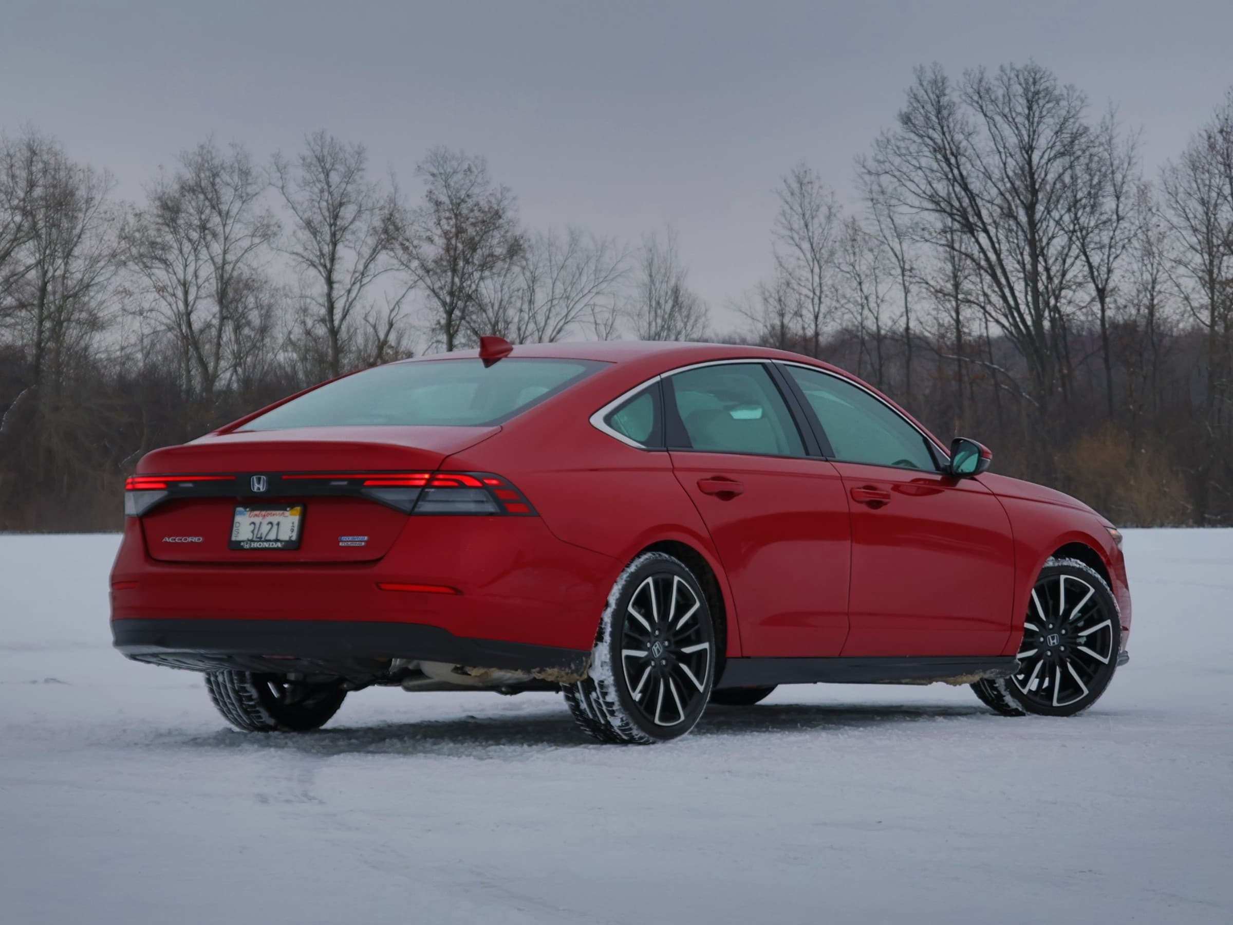 Rear three-quarter view of a red Honda Accord Hybrid sedan with black and silver wheels, parked on a snowy field in front of bare trees.
