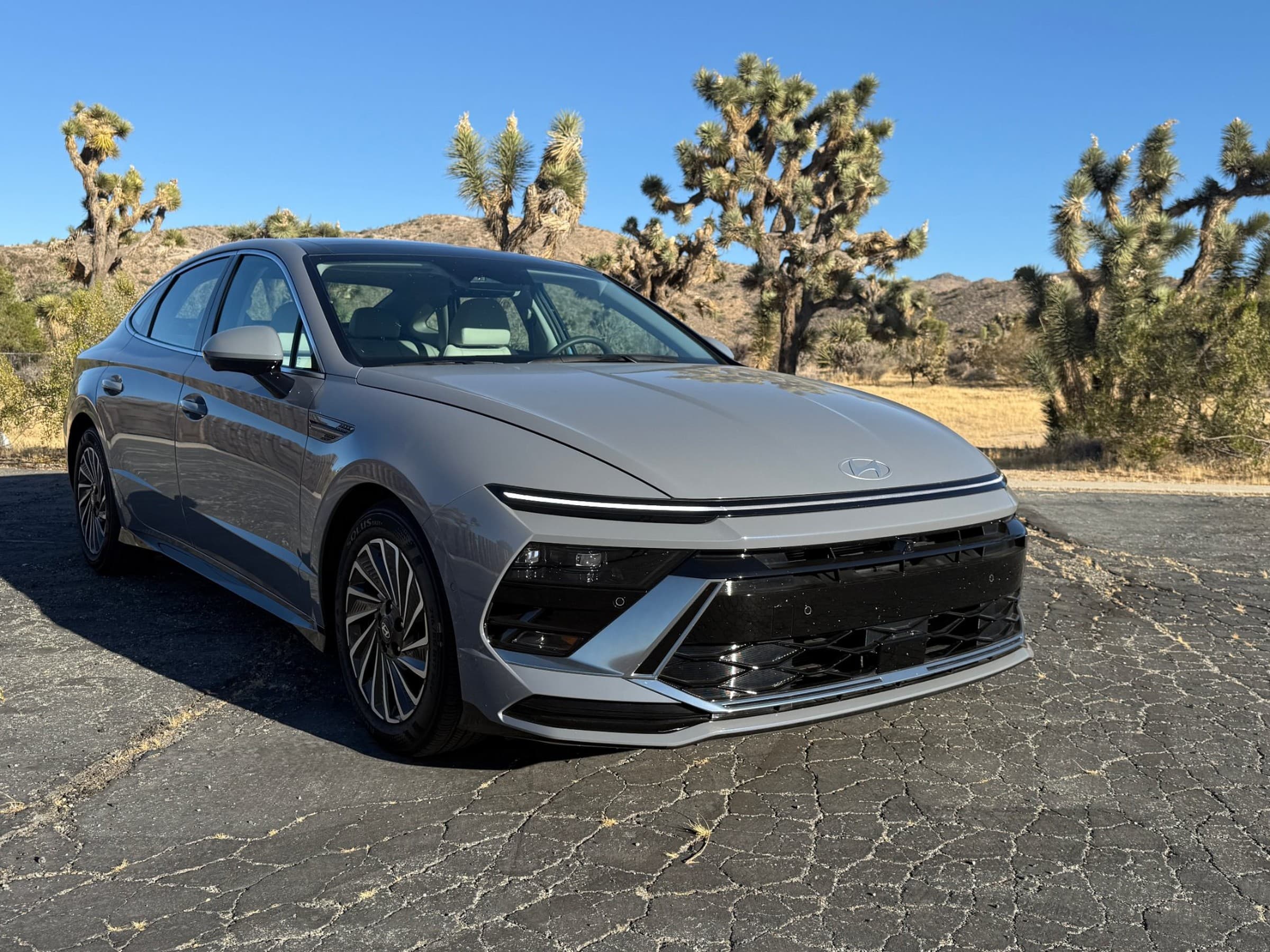Front three-quarter view of a gray 2026 Hyundai Sonata Hybrid with a full-width front light bar, parked on cracked pavement among Joshua trees.
