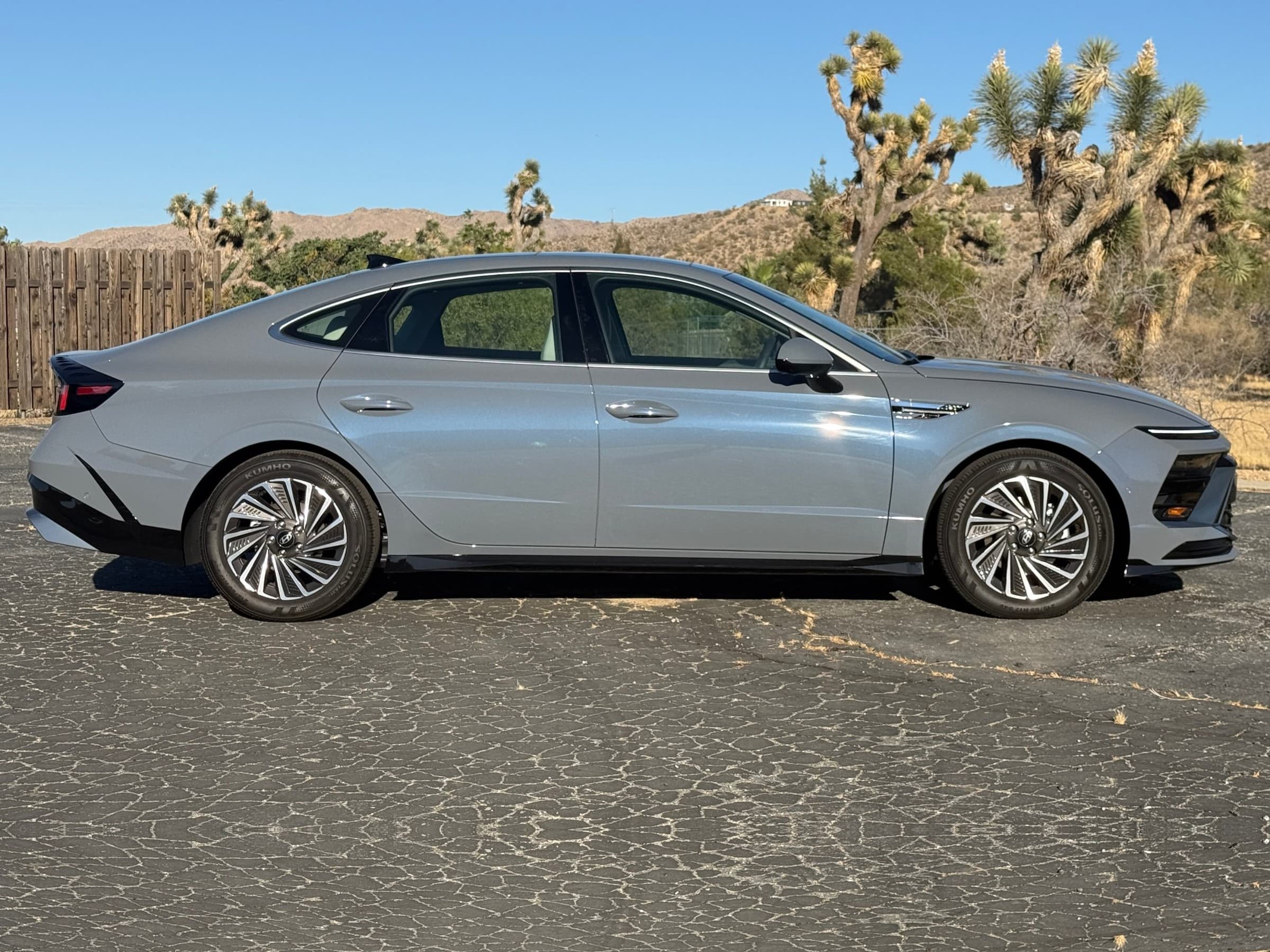 Side profile of a gray Hyundai Sonata Hybrid with turbine-style wheels, parked on pavement with Joshua trees and desert hills behind.