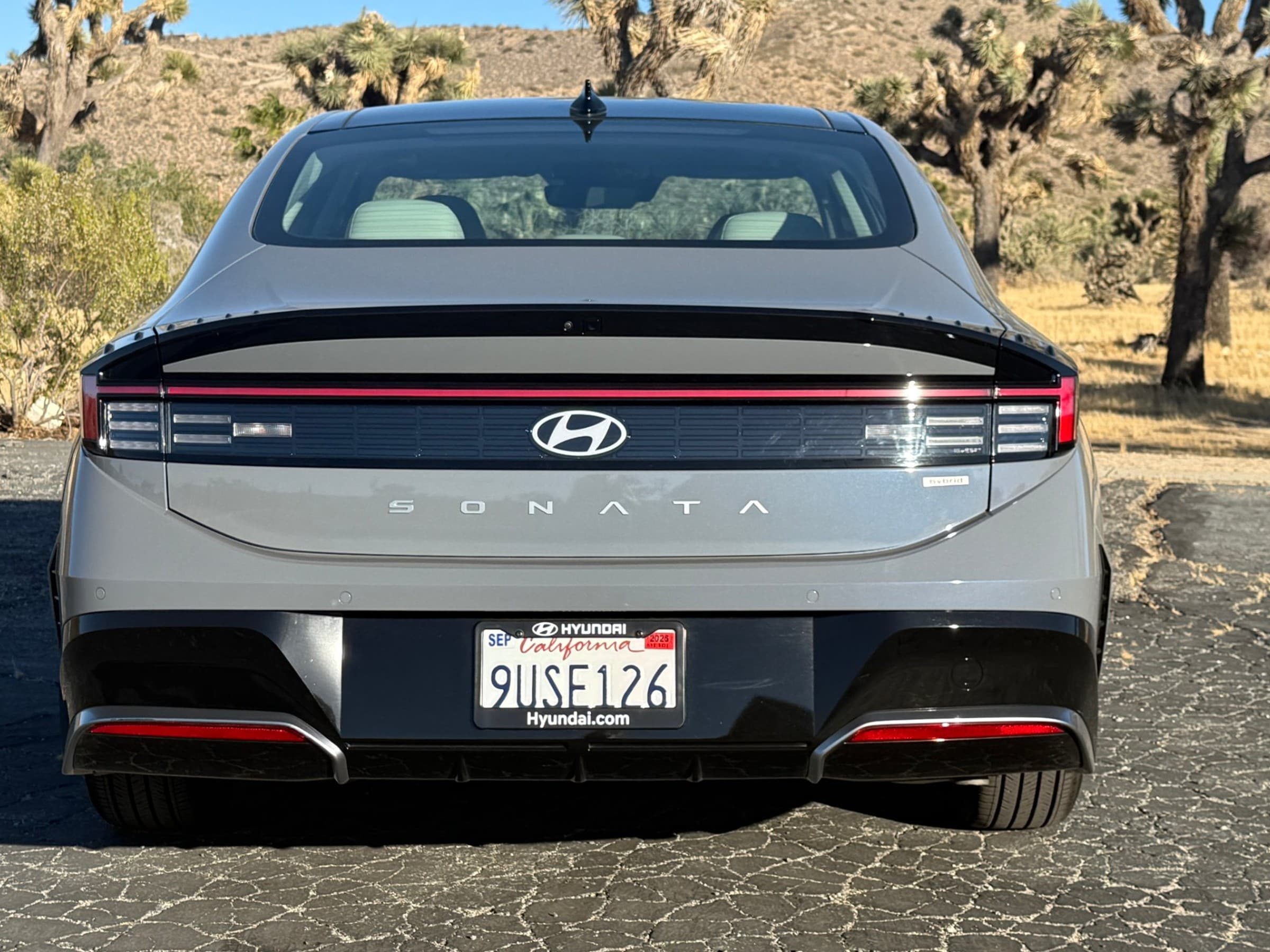 Straight-on rear view of a gray Hyundai Sonata Hybrid with a full-width red light bar, Hyundai logo and Sonata lettering, with Joshua trees behind.