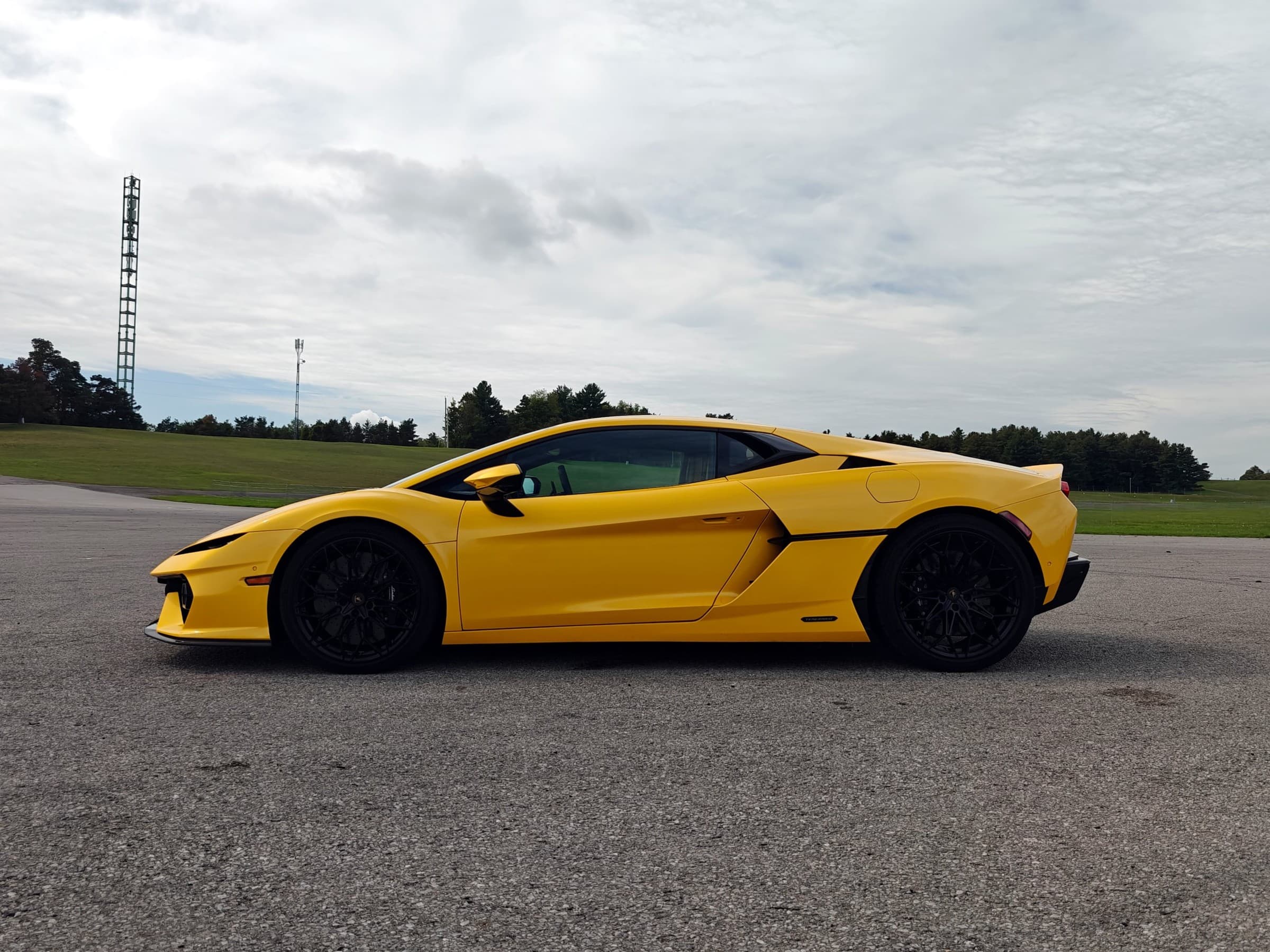 Yellow 2026 Lamborghini Temerario parked on open tarmac at a racetrack, seen from the front driver’s side under a cloudy sky.