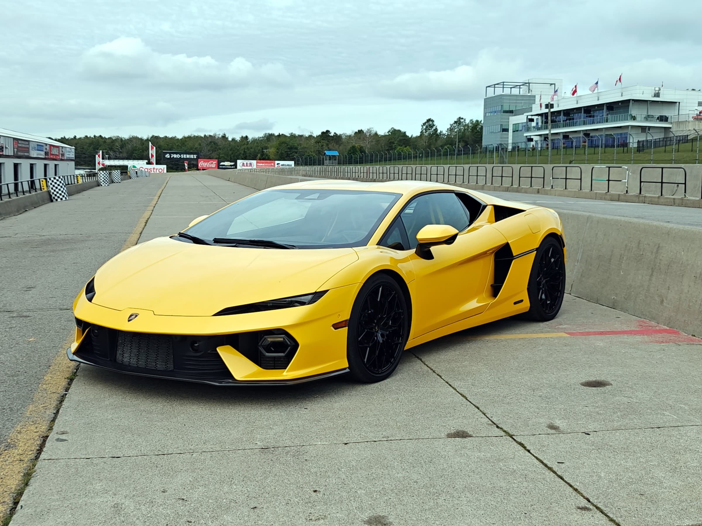 Yellow 2026 Lamborghini Temerario parked in a racetrack pit lane, seen from the front driver’s side, with black wheels and the grandstand behind.