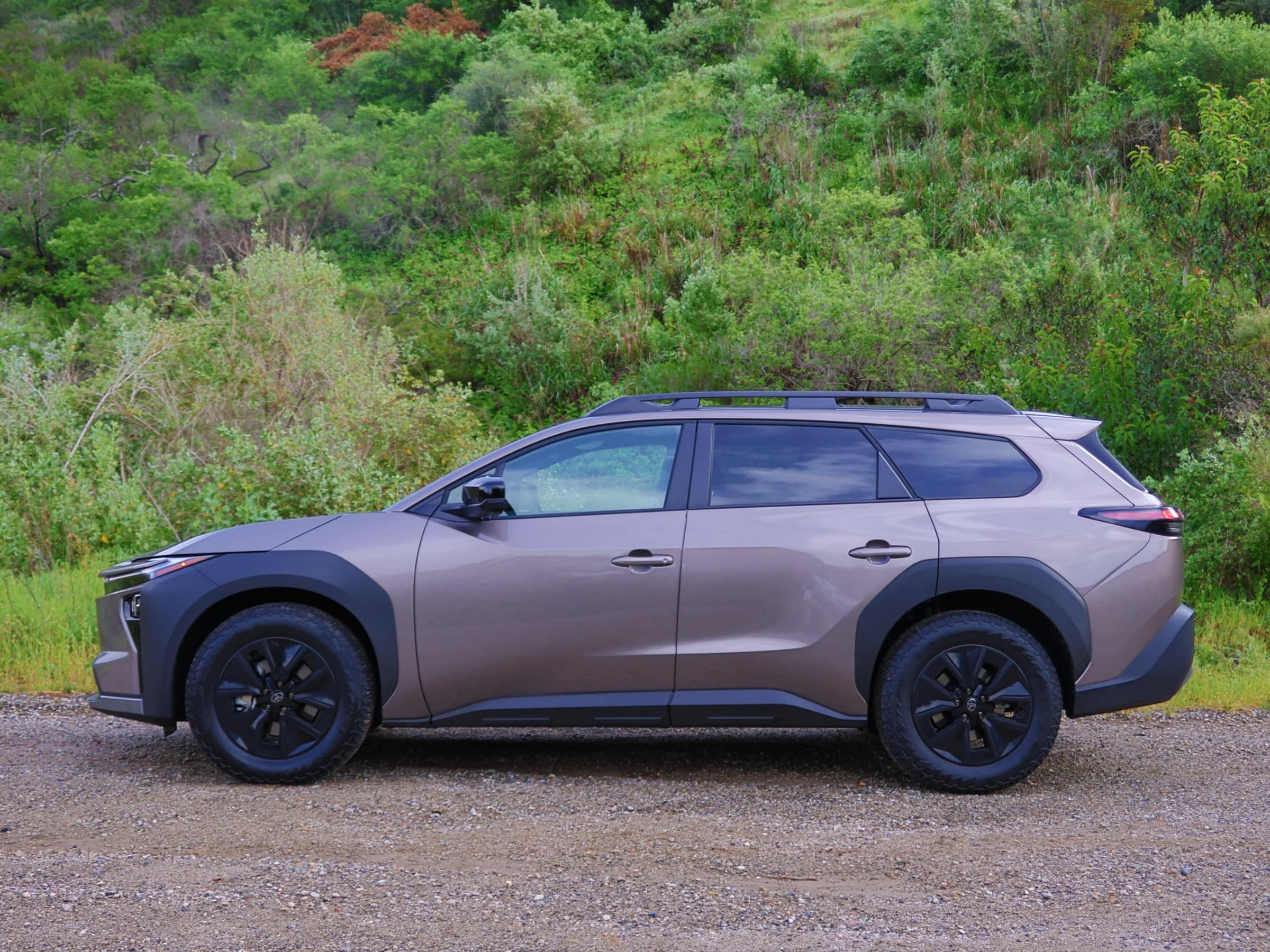 Side profile of a bronze Toyota bZ Woodland showing its wagon-like shape, roof rails, black fender flares and all-terrain tires, against a brushy hillside.