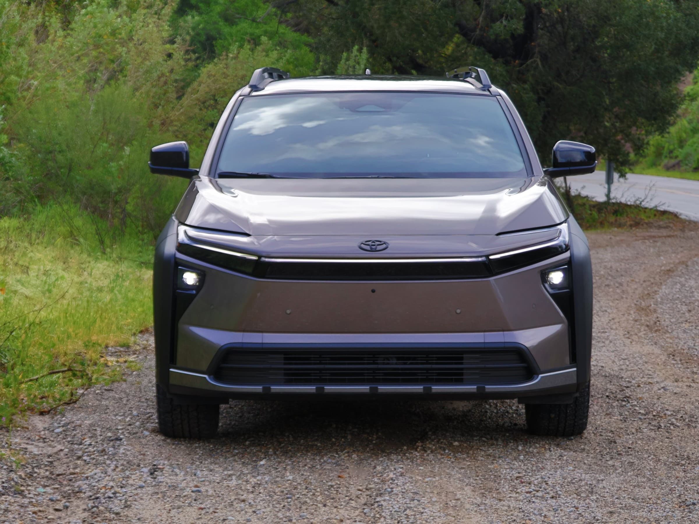 Head-on view of a bronze Toyota bZ Woodland with its lights on, parked on a gravel shoulder beside a winding road.