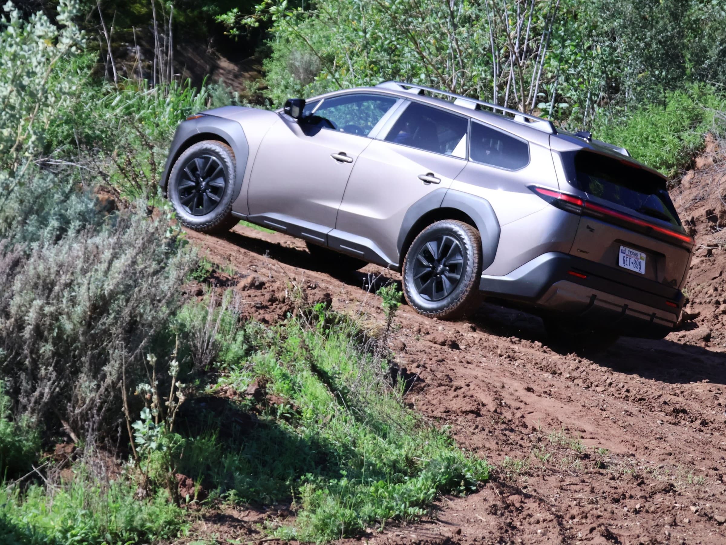 Bronze Toyota bZ Woodland climbing a steep dirt trail between brushy banks, seen from the rear driver’s side.