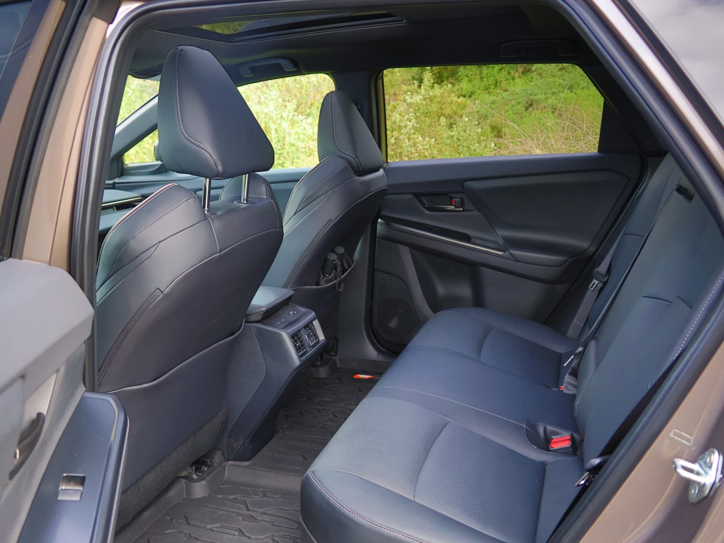 Rear seat of a Toyota bZ Woodland seen through the open back door, with black upholstery, rear air vents, all-weather floor mats and a sunroof overhead.
