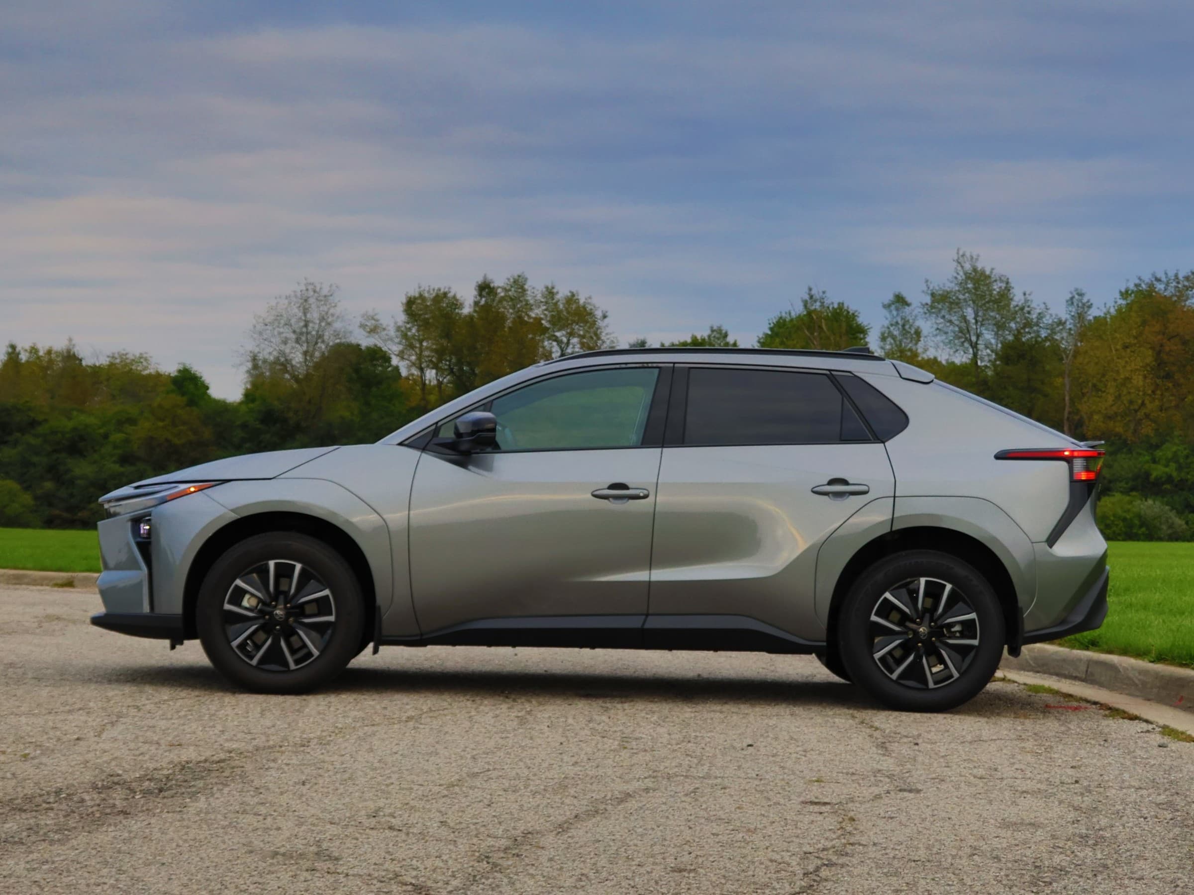 Side profile of a gray Toyota bZ crossover with a black roof rail and two-tone wheels, parked on a curved lot with trees behind.
