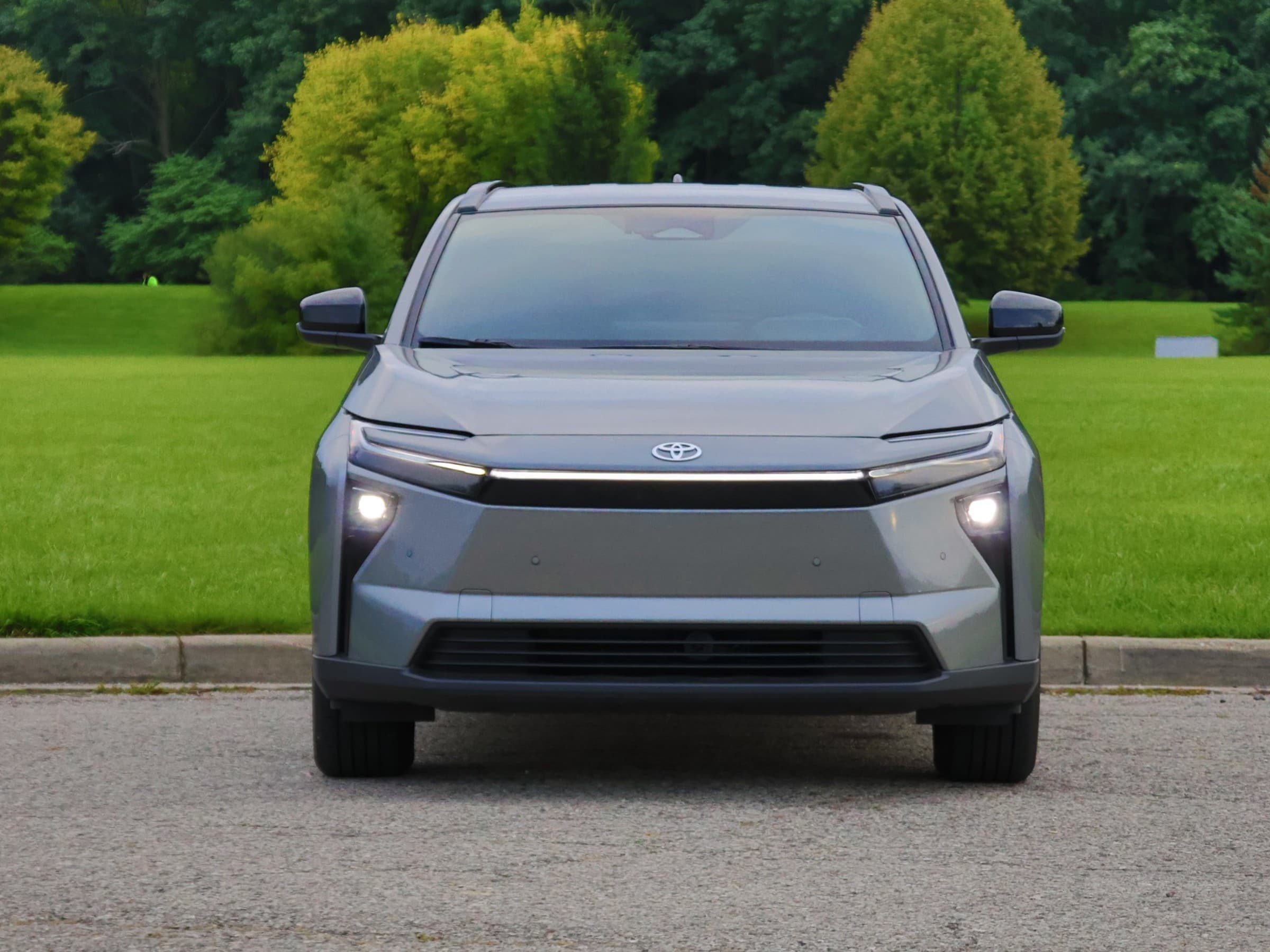 Head-on front view of a gray Toyota bZ with its headlights on, showing the full-width light bar, Toyota logo and smooth body-colored front panel.