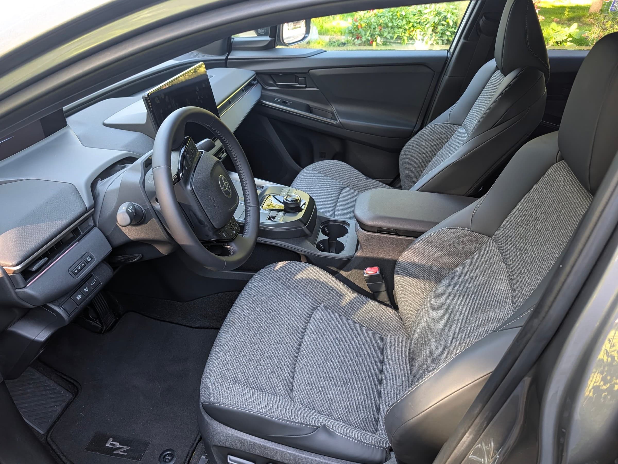 Front seats of a Toyota bZ seen through the open driver’s door, with gray fabric upholstery, a floating center console and a bZ logo on the floor mat.