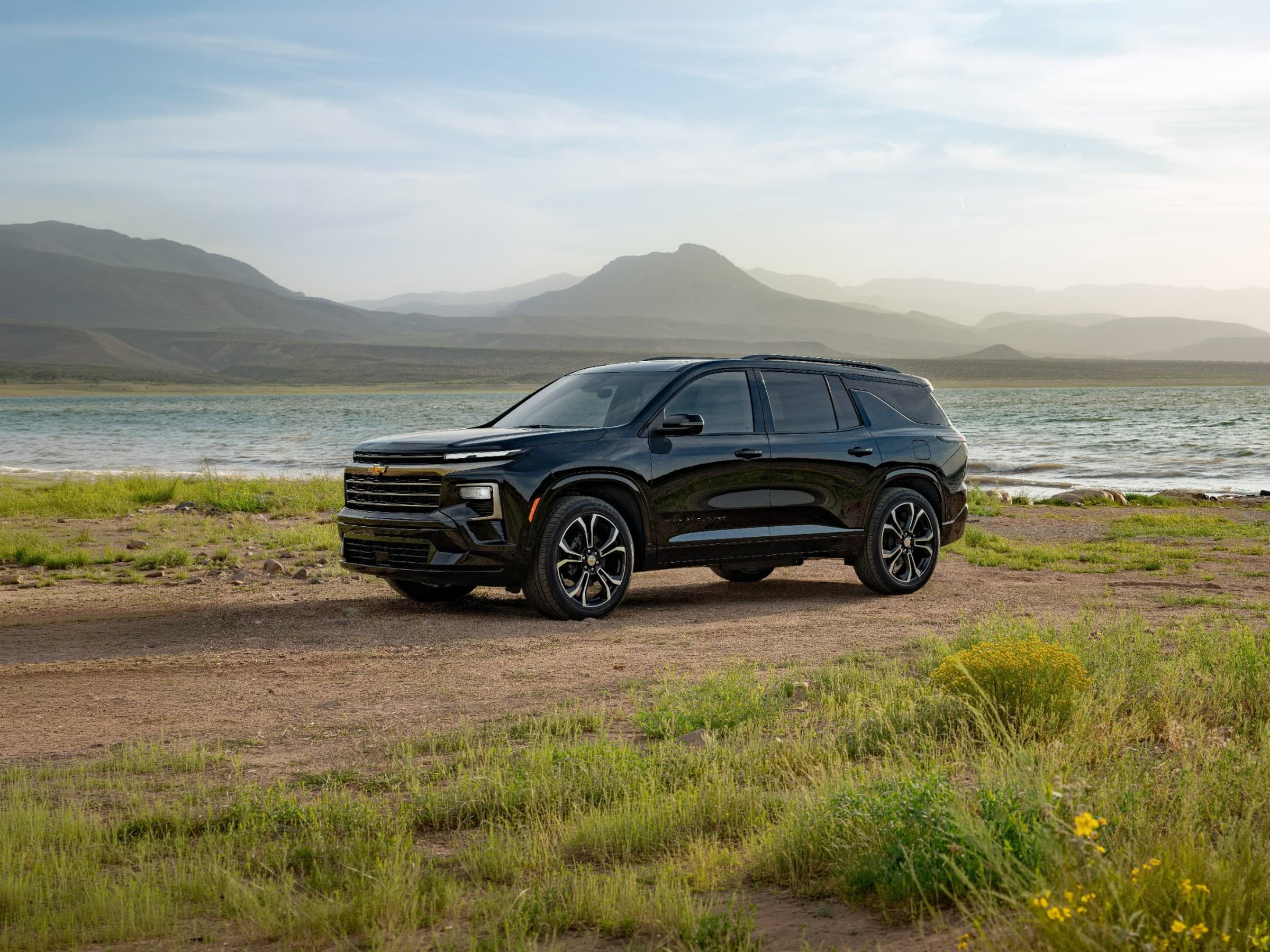 Front three-quarter view of a black 2027 Chevrolet Traverse High Country with two-tone wheels, parked on a dirt shore beside a lake with hazy mountains behind.