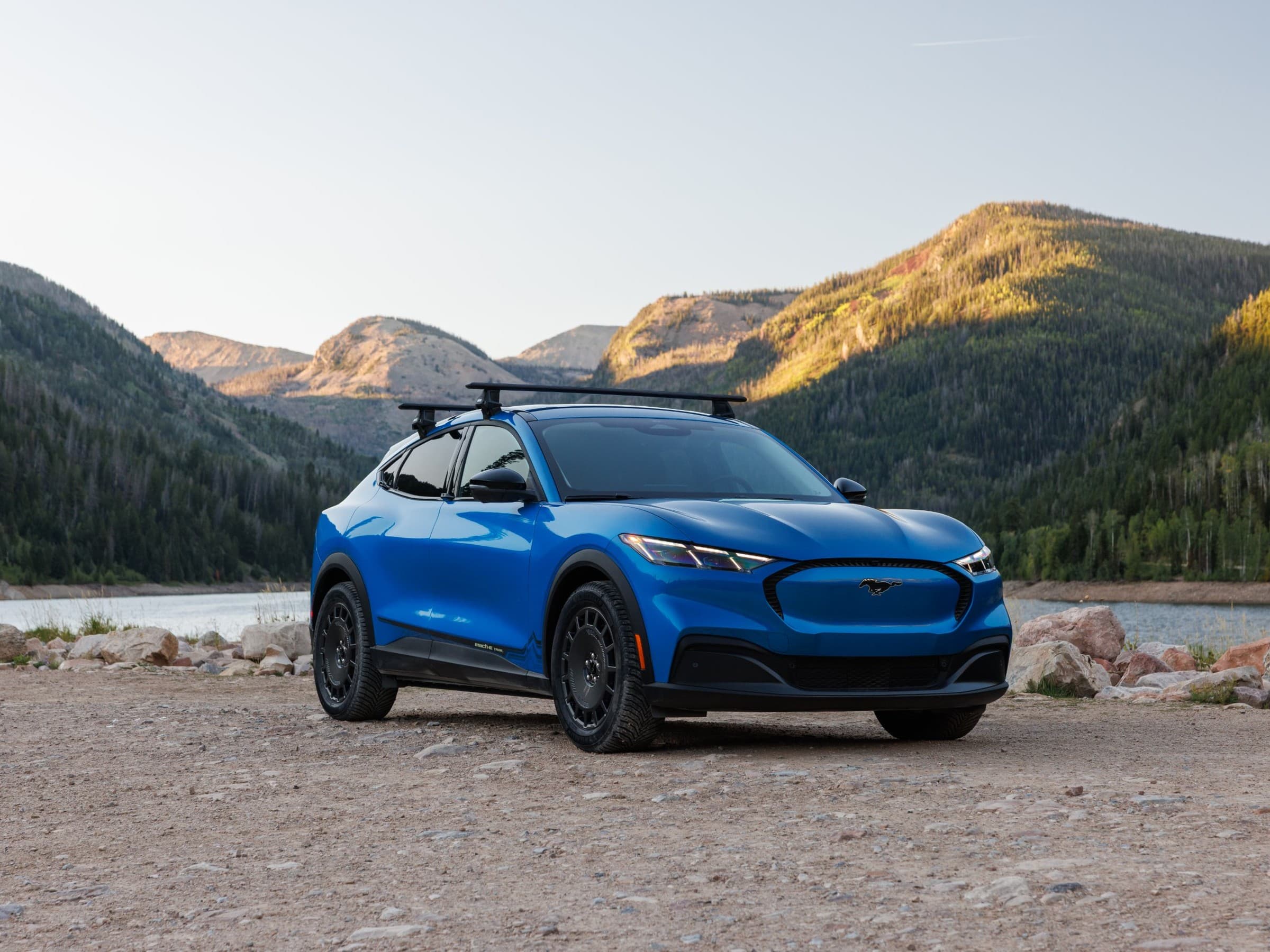 Front three-quarter view of a blue Ford Mustang Mach-E Trail with black wheels, all-terrain tires and roof crossbars, parked by a mountain lake at sunset.