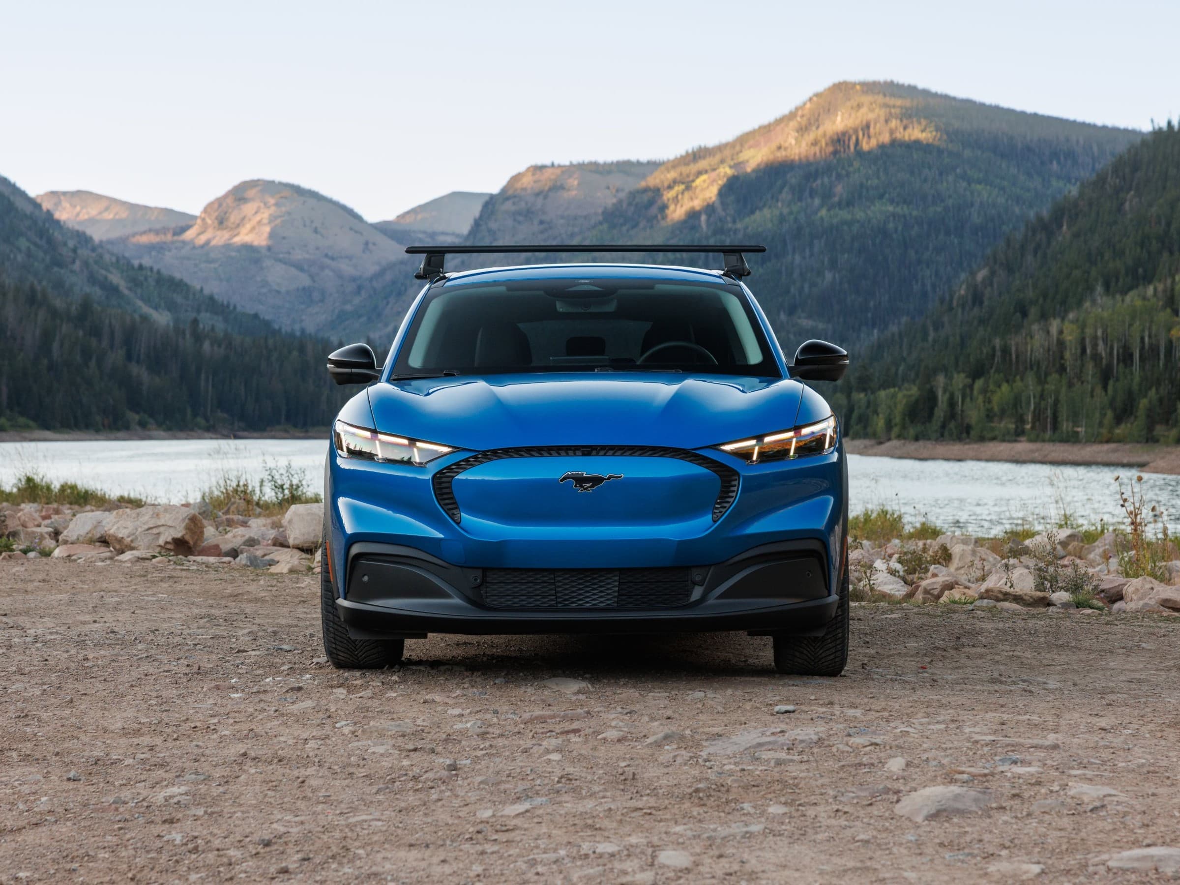 Head-on view of a blue 2027 Ford Mustang Mach-E Trail with a black roof rack, parked on a gravel shore with a lake and forested mountains behind.