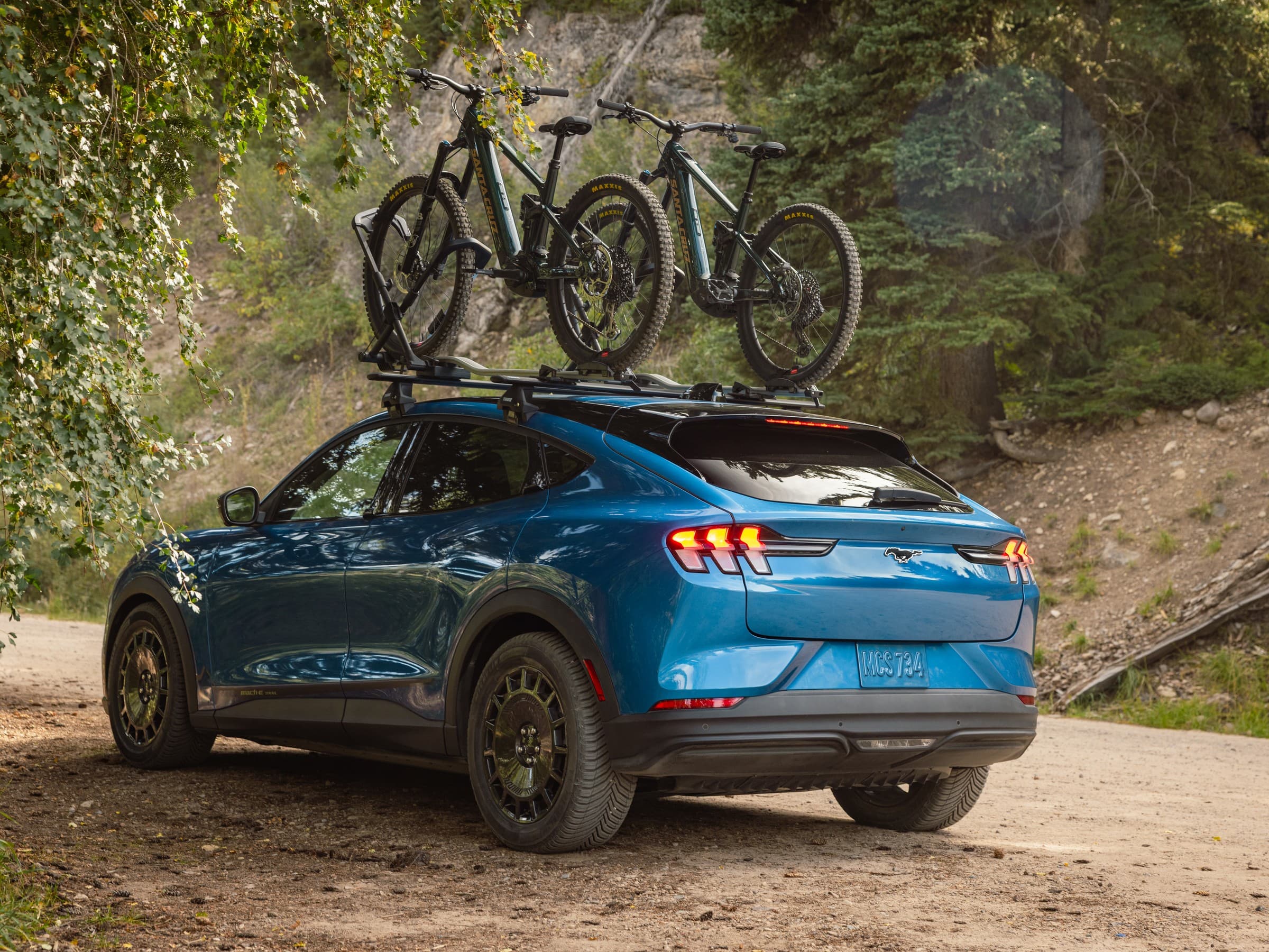 Rear three-quarter view of a blue Ford Mustang Mach-E Trail carrying two mountain bikes on a roof rack, parked on a dirt road beside a wooded slope.