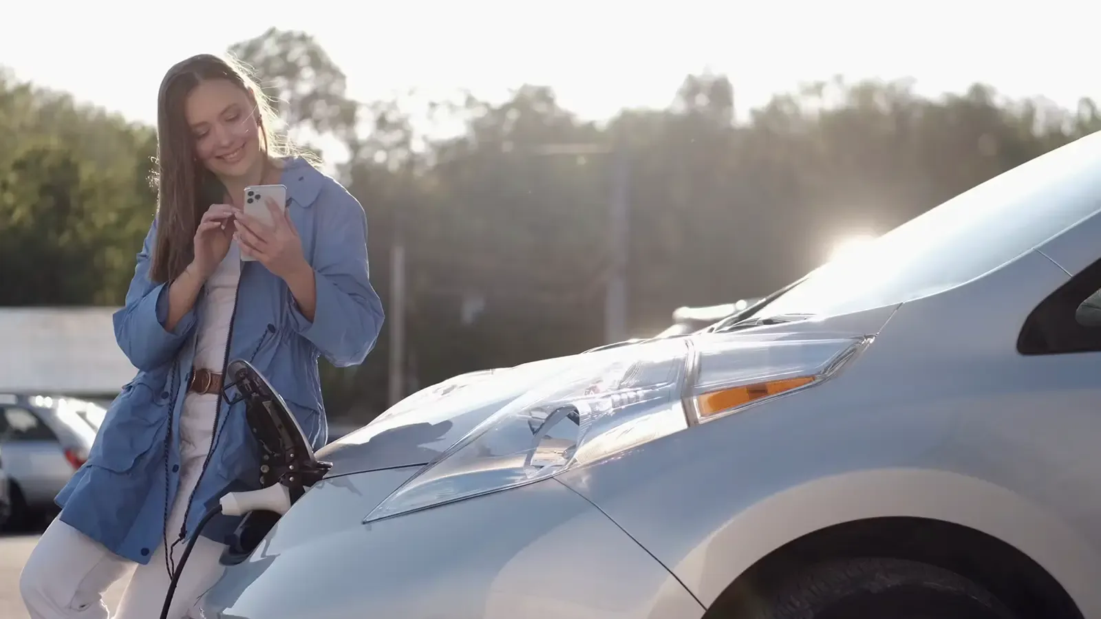 Woman standing, using her phone, and happily glancing at her car charging