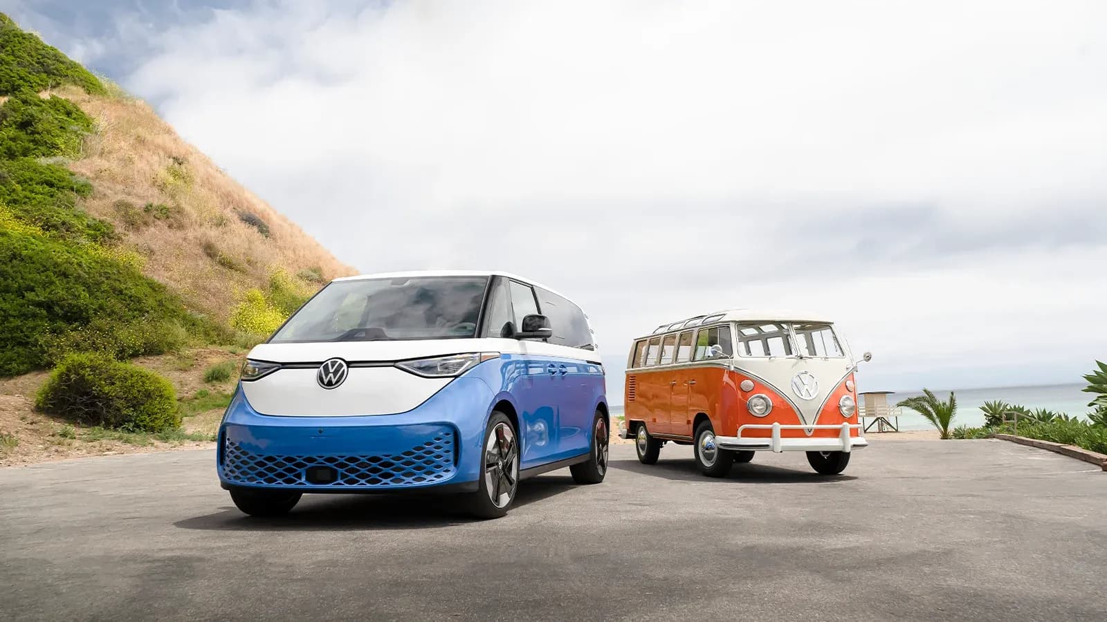a blue ID.Buzz next to an orange, classic Volkswagen buss parked at an ocean viewpoint next to a grass-covered hillside