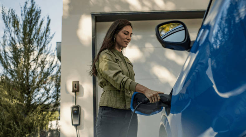 Una persona conecta un cable de carga a un vehículo eléctrico azul junto a un garaje.