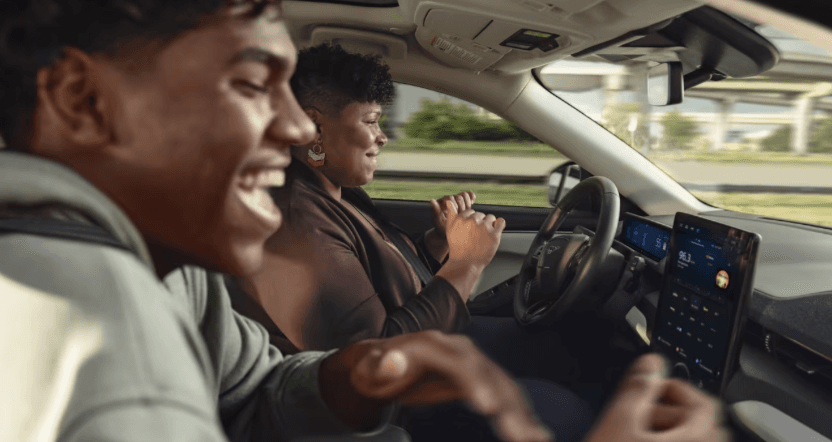 Dos personas viajan dentro de un auto; una persona conduce y la otra sonríe en el asiento delantero.