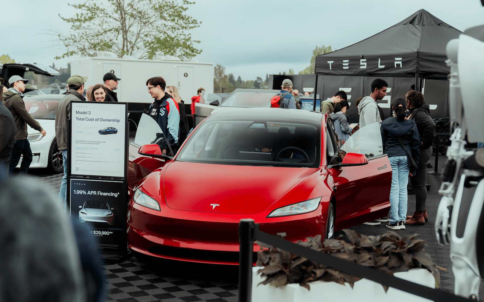 Front view of a red electric sedan with open doors at a Tesla display, with people gathered around.