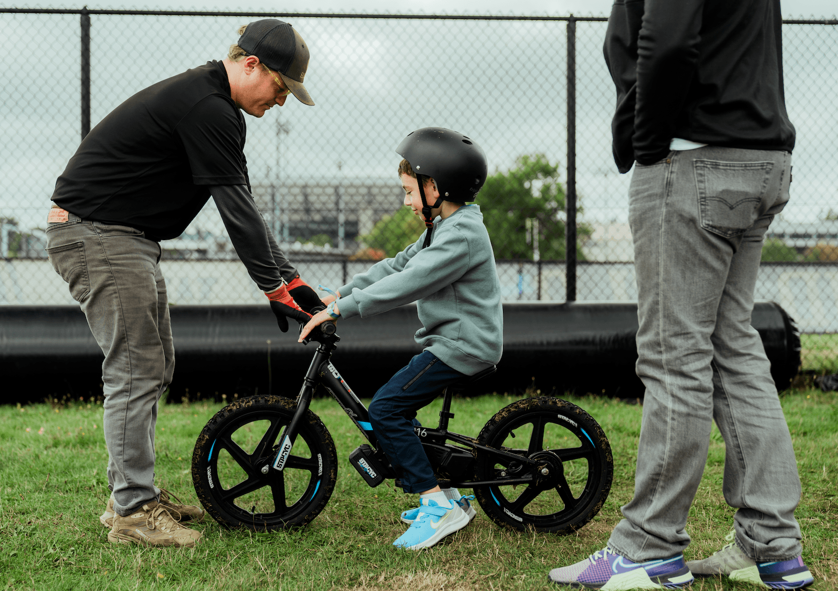 A person helps a child balance on a small bike wearing a helmet on grass.