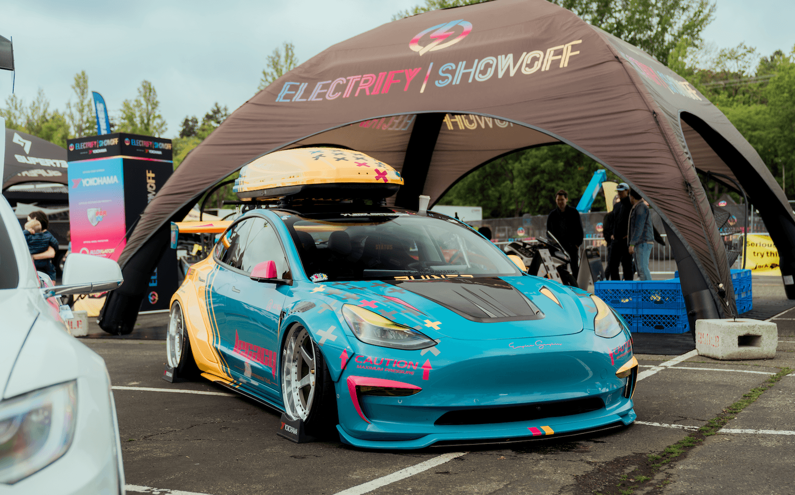 A customized blue electric sedan with a roof cargo box sits under an Electrify Showoff tent.