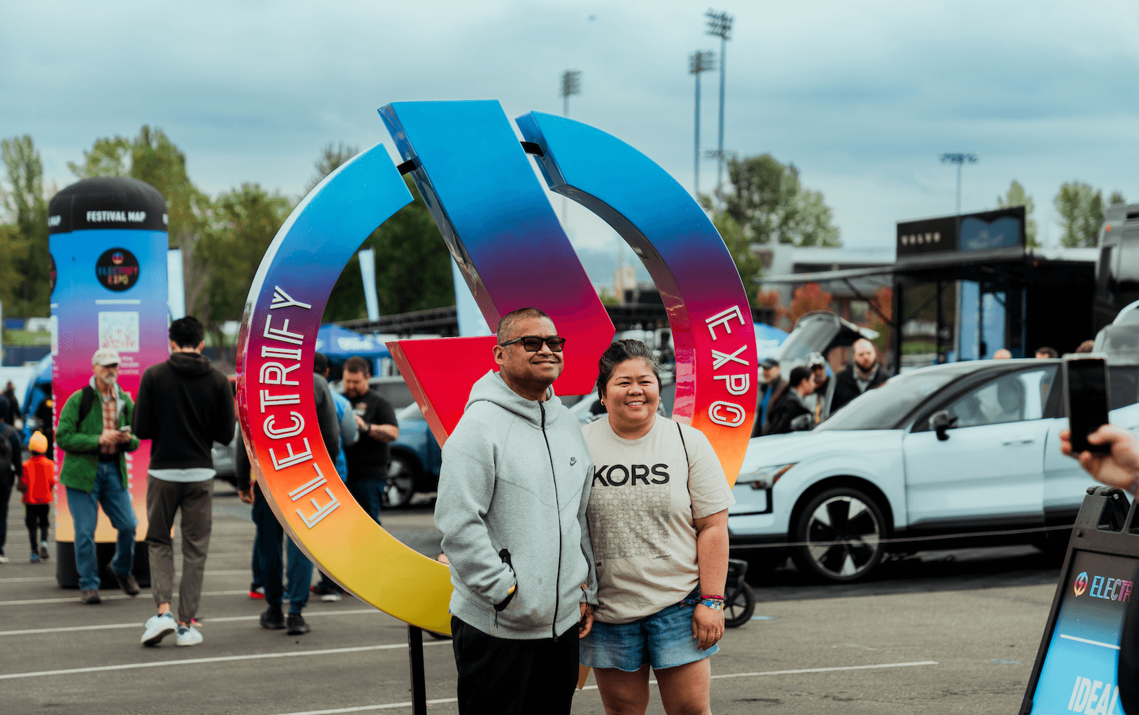 Two people pose in front of a colorful Electrify Expo sign at an outdoor event.