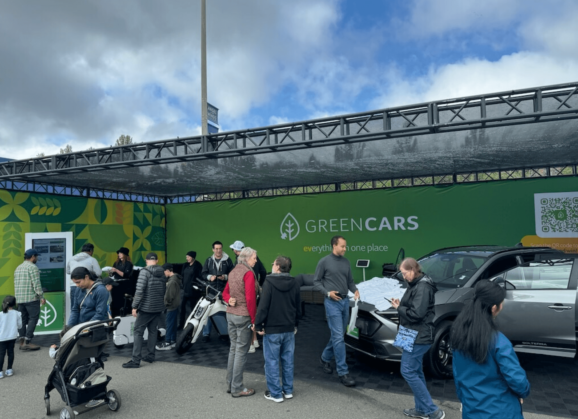 People gather around a GreenCars booth beside a dark SUV and a stroller.