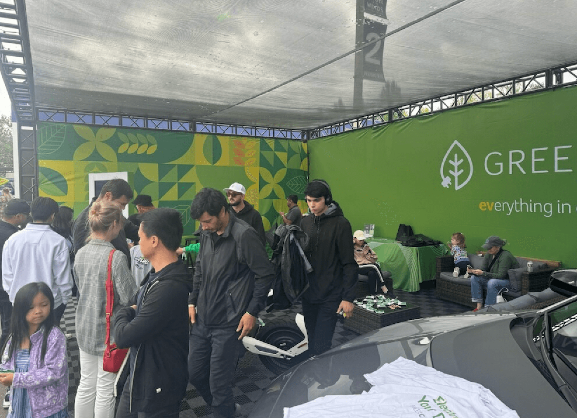 People gather under a green GreenCars booth beside a gray electric sports car.