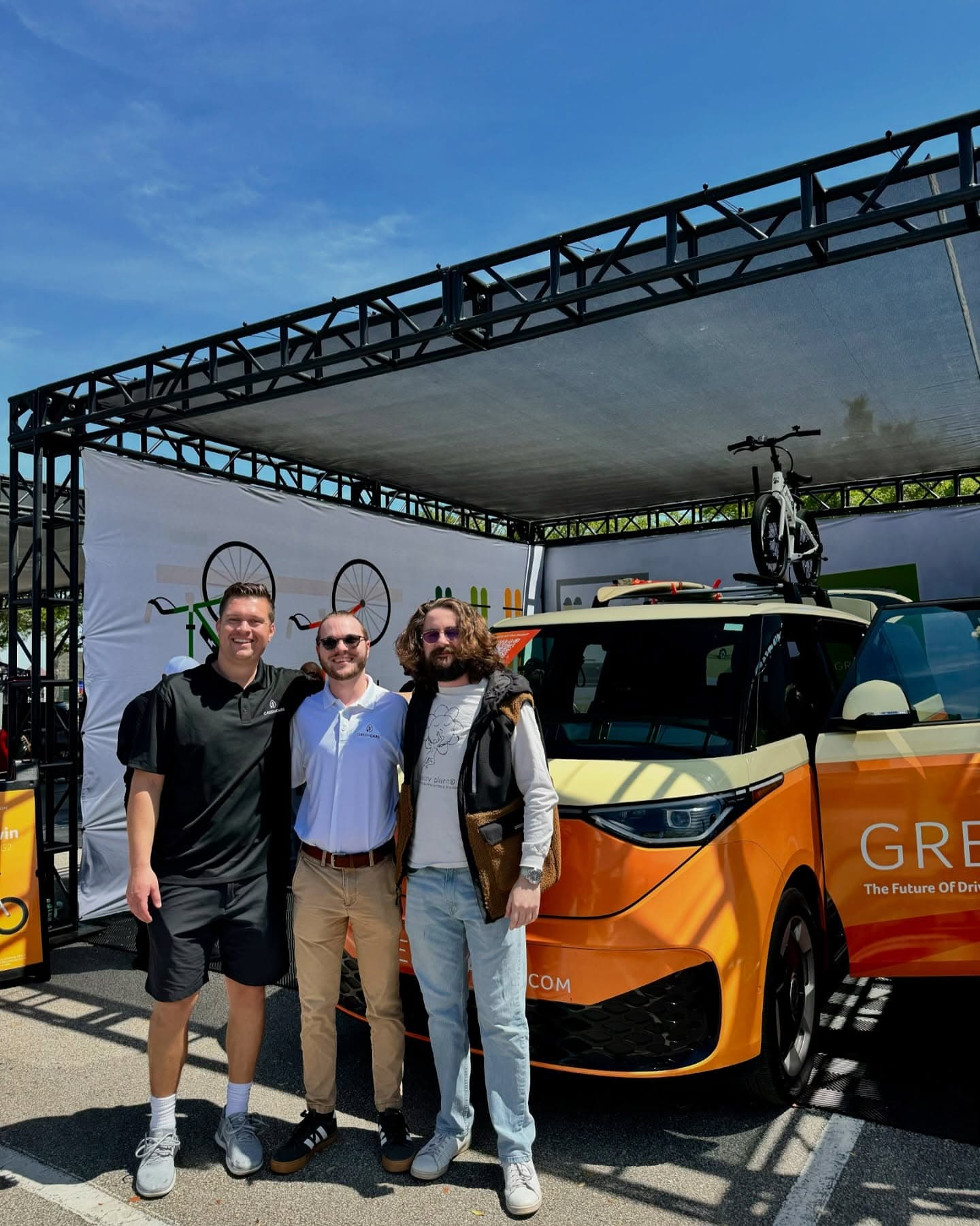 Three people stand beside a GreenCars electric bus with a bicycle mounted on top.