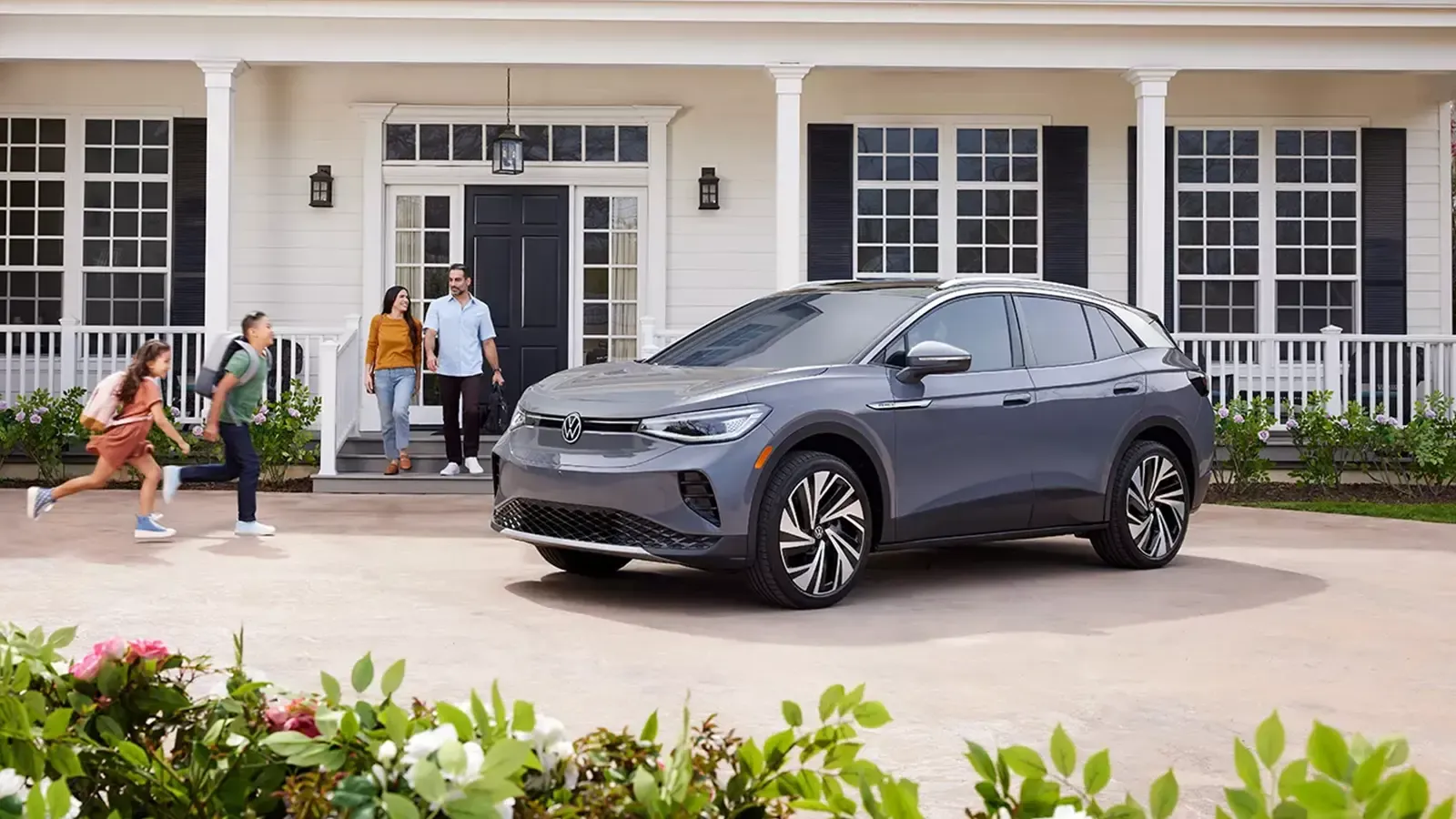 A gray Volkswagen ID.4 is parked in a driveway as two adults stand by the front door and two children run past.