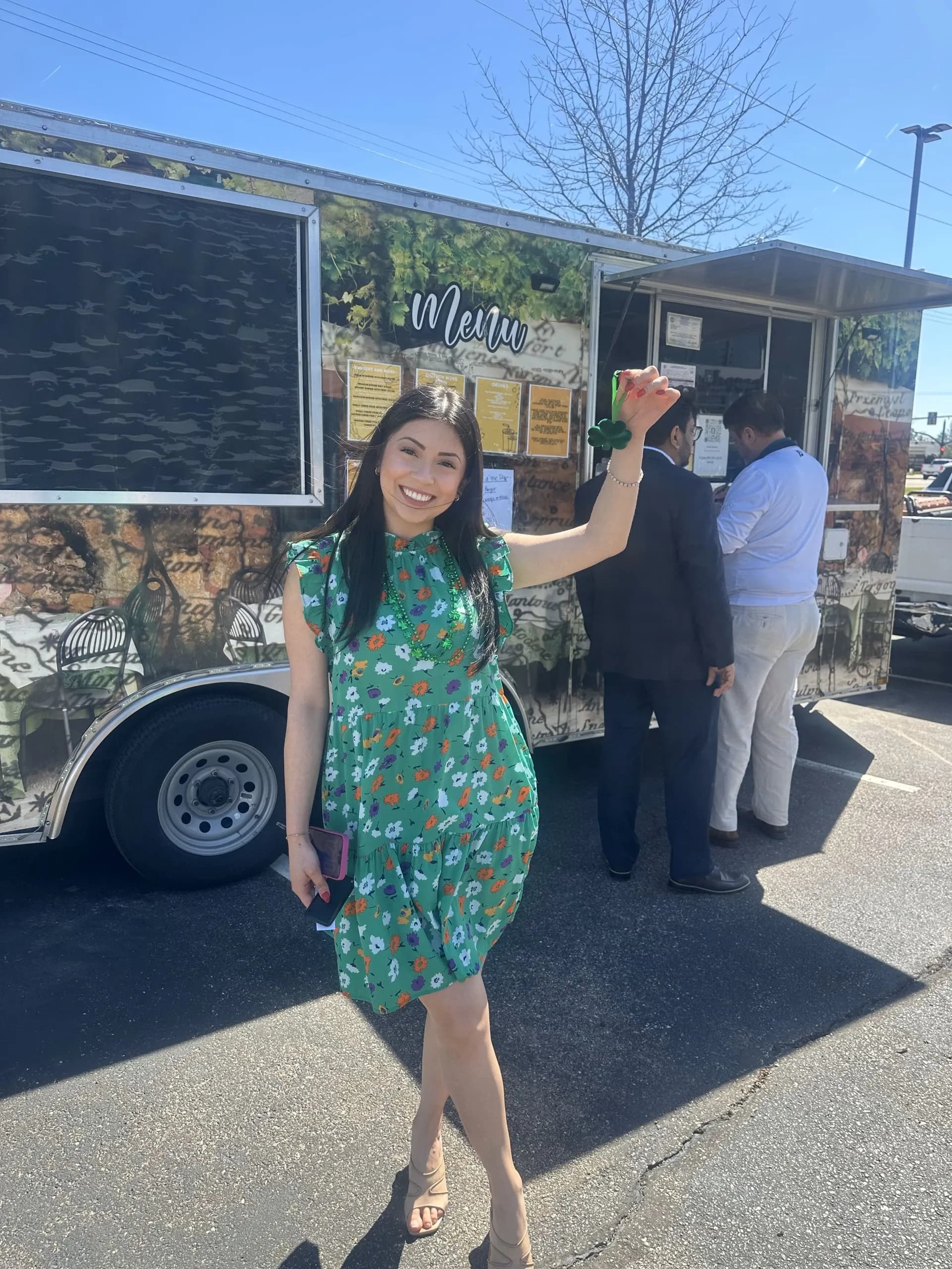 Una persona sonriente con un vestido verde está junto a un tráiler de comida mientras dos personas hacen un pedido.
