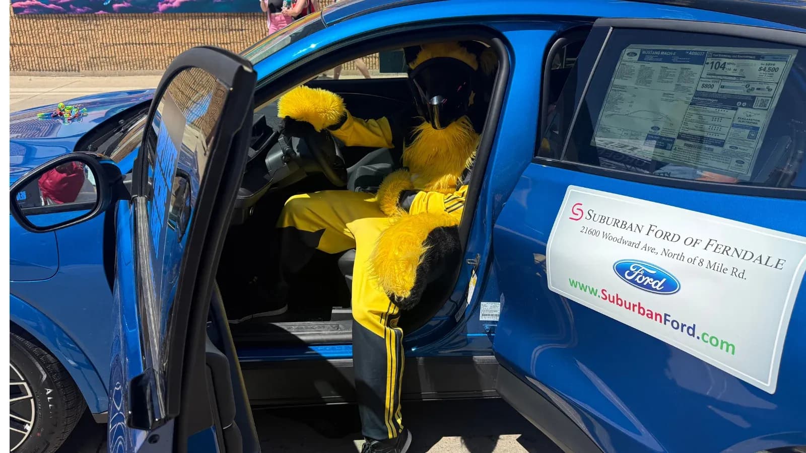 Open side view of a blue Ford Mustang Mach-E with a person in a yellow costume sitting in the driver’s seat.