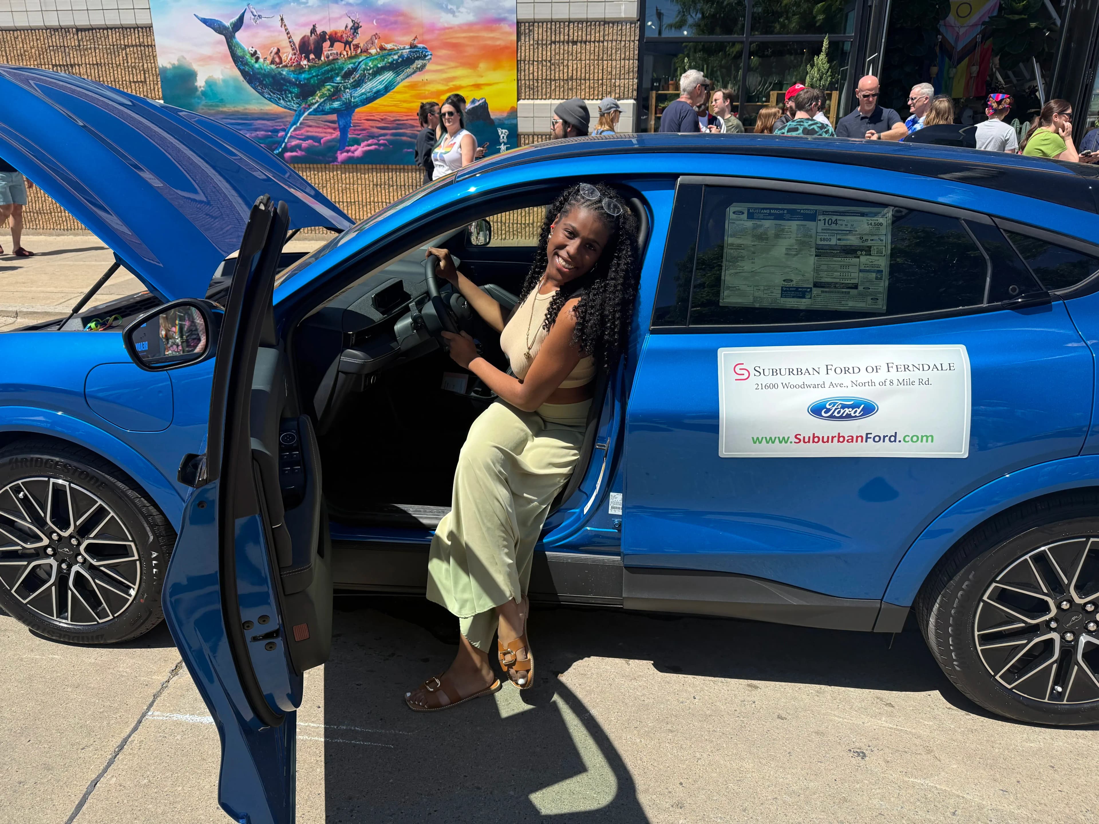 A person sits in the driver’s seat of a blue Ford Mustang Mach-E with the door open at a Pride event.