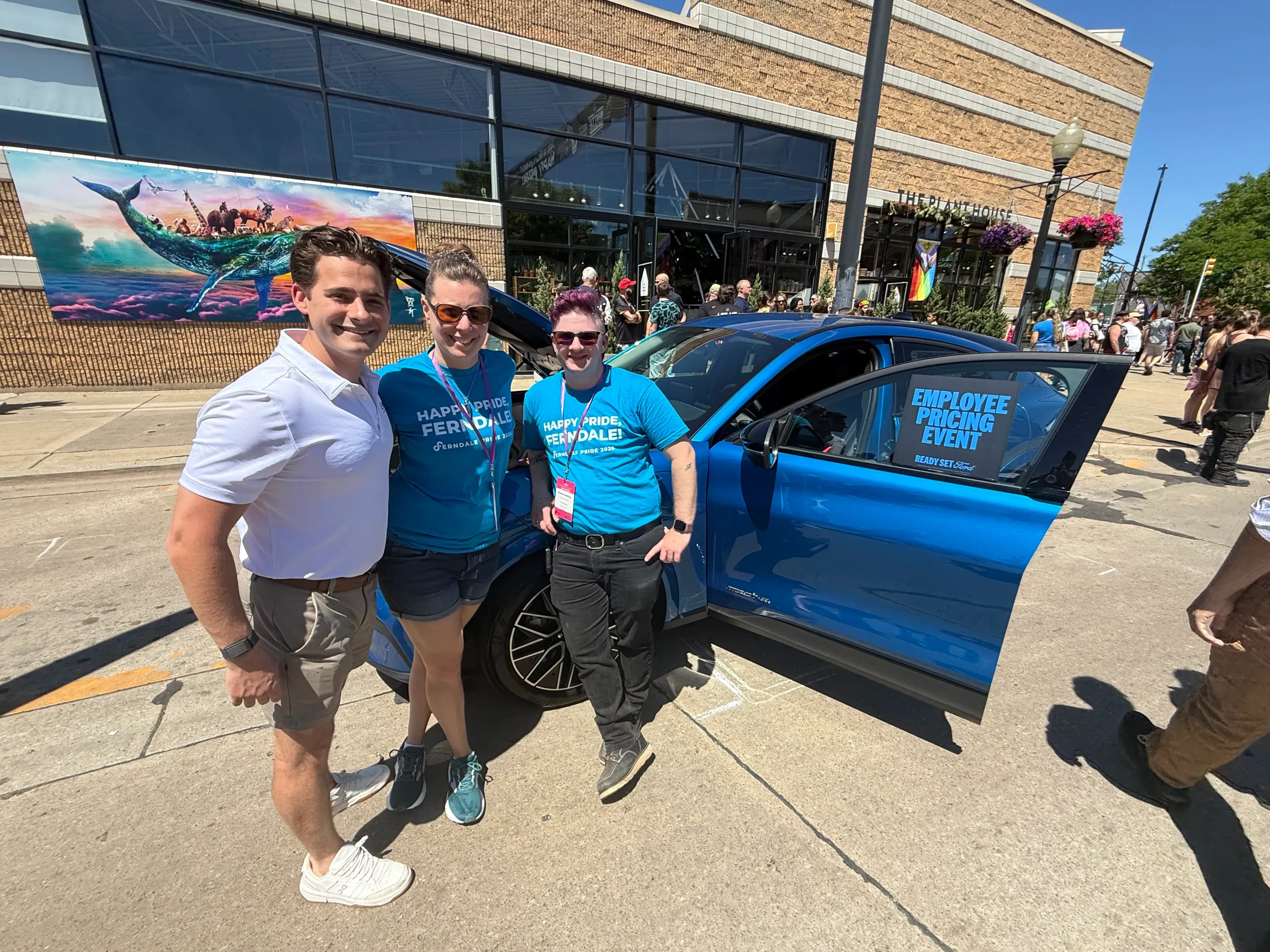 Three people stand beside a blue Ford Mustang Mach-E with its door open at a Pride event.