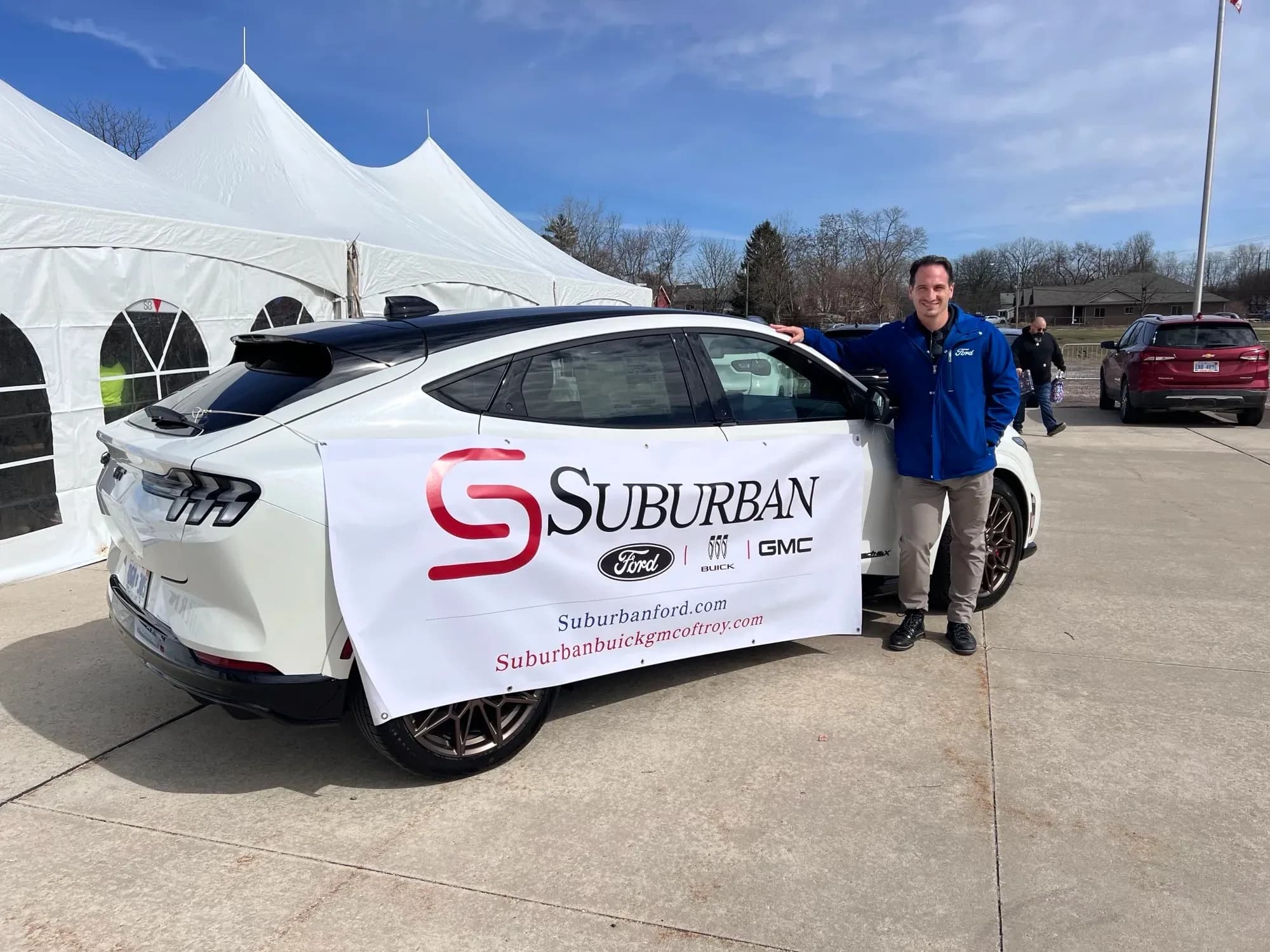 Side view of a white Ford Mustang Mach-E with a Suburban banner and a person standing beside it.