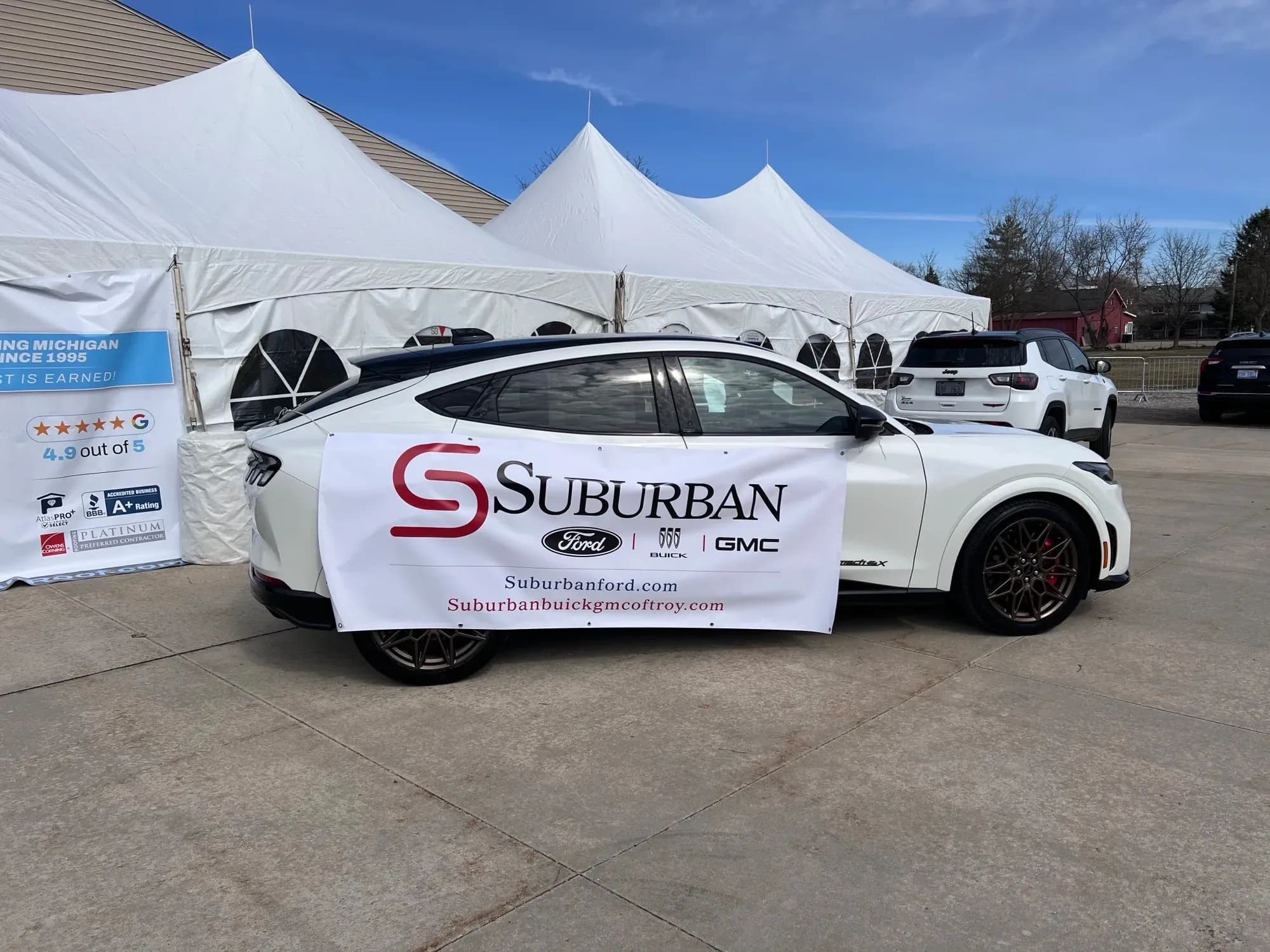 Side view of a white Ford Mustang Mach-E with a Suburban banner beside event tents.