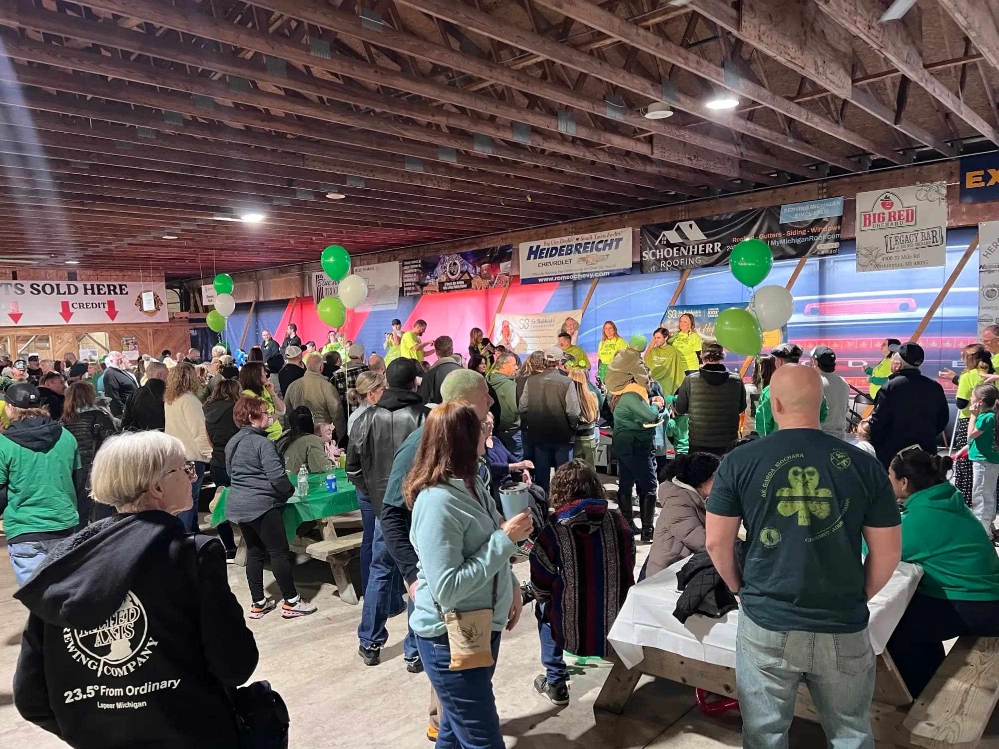 Crowd gathered inside a barn-like venue with people on stage and green balloons during a St. Baldrick’s event.