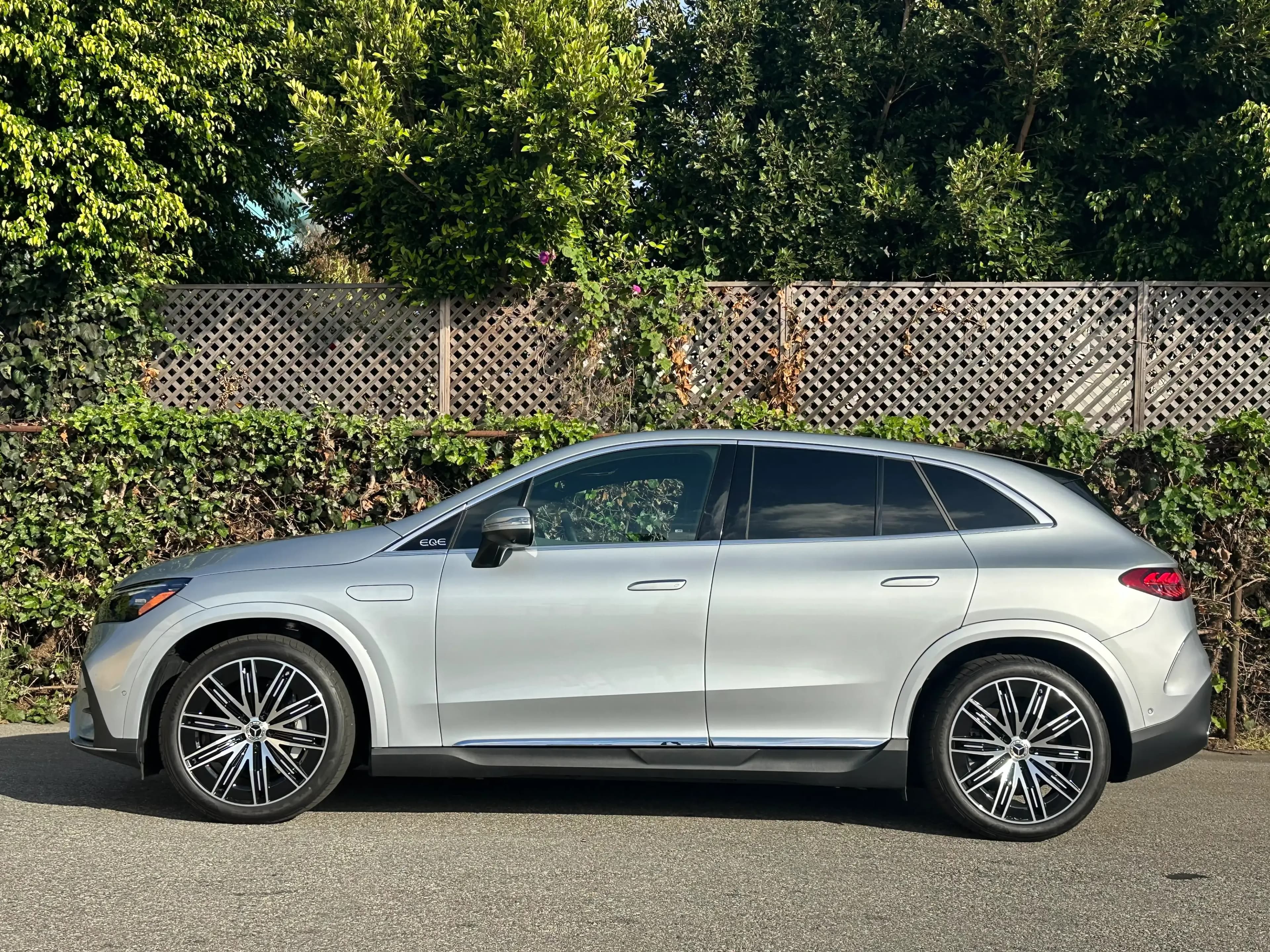 Side profile of a silver Mercedes-Benz EQE SUV parked beside a fence and trees.