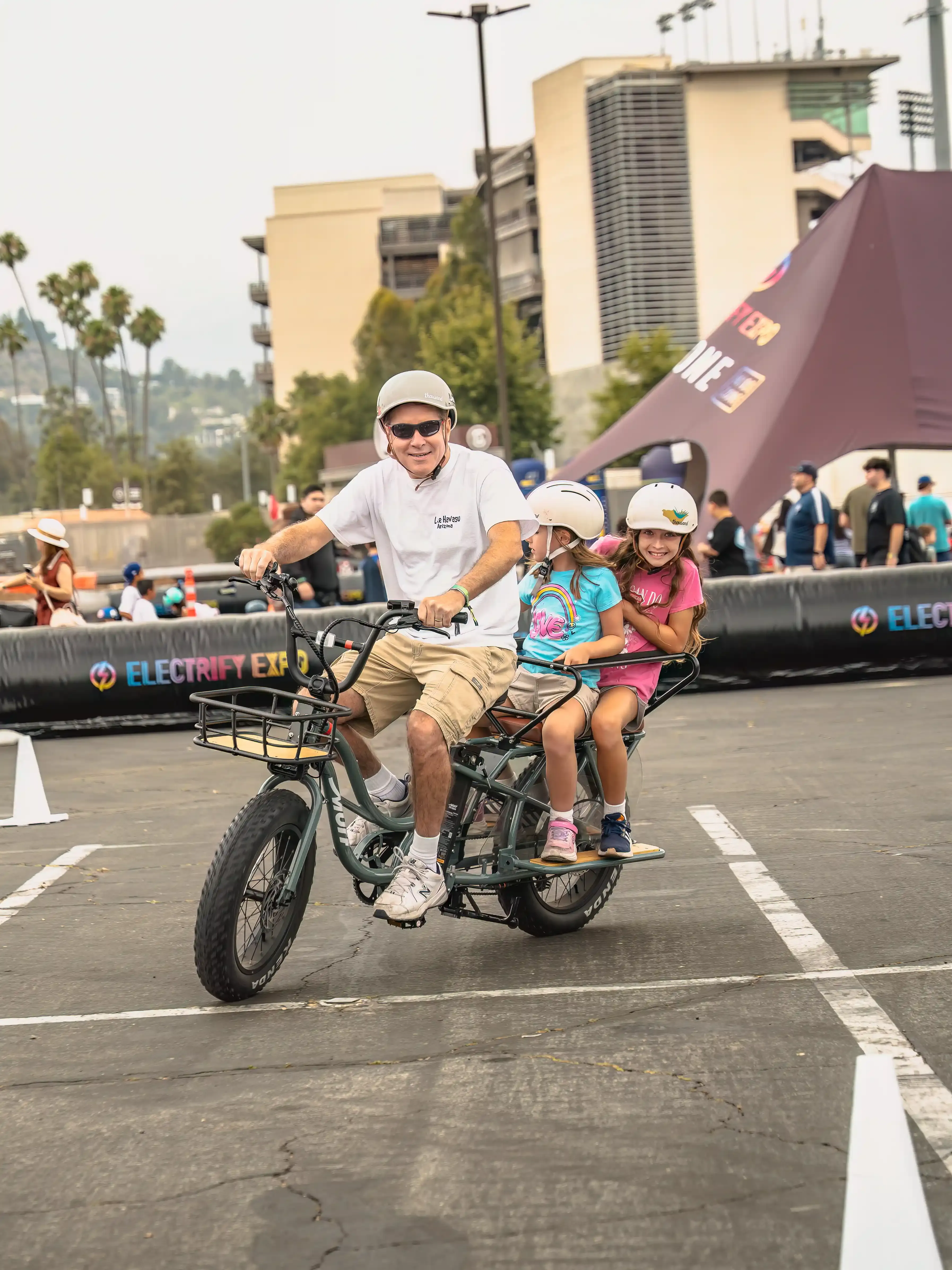 Un hombre y dos niños viajan en una bicicleta eléctrica de carga larga en un evento al aire libre.