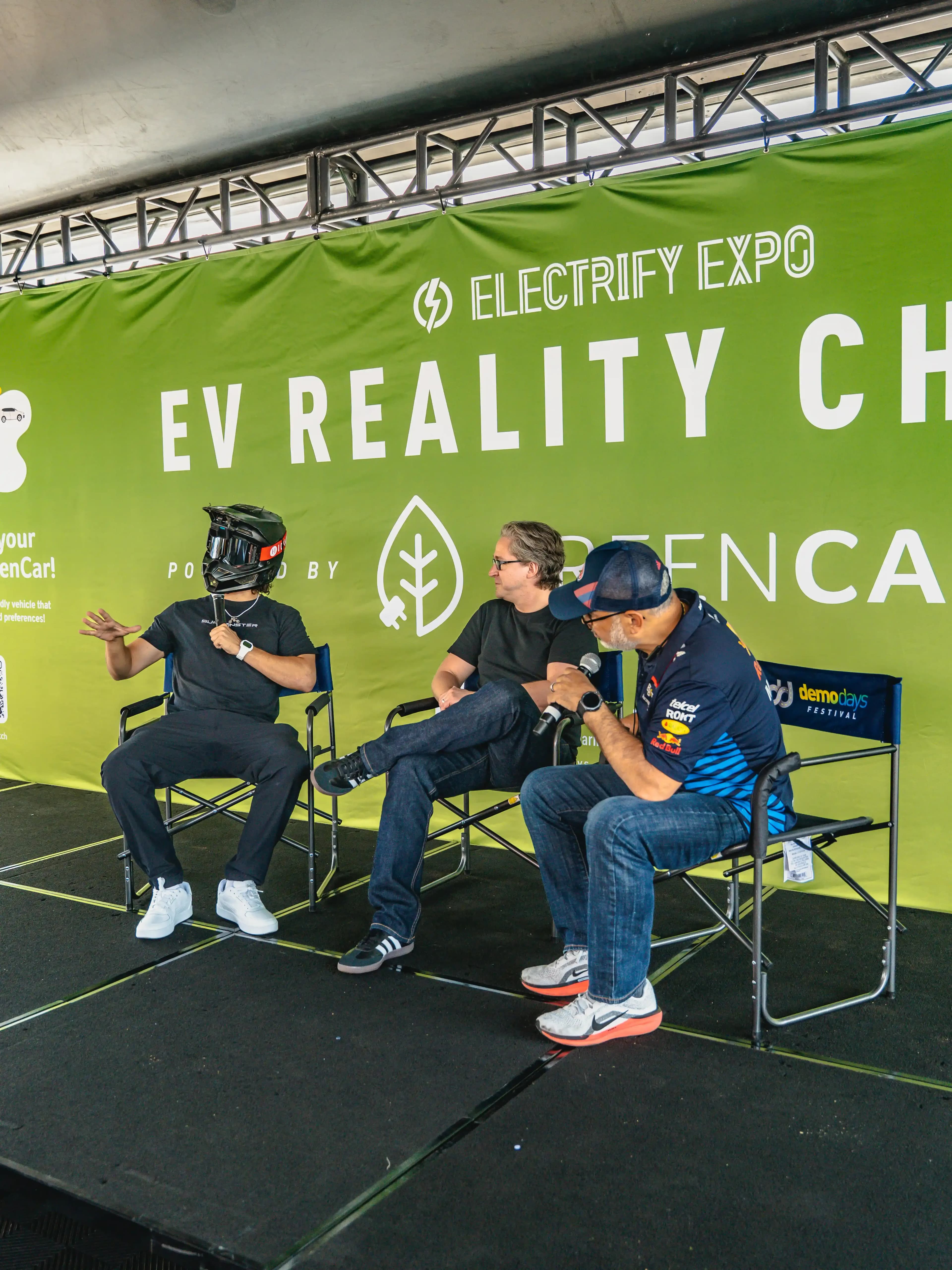 Tres personas se sientan en un escenario frente a un letrero de Electrify Expo EV Reality Check; una lleva un casco.