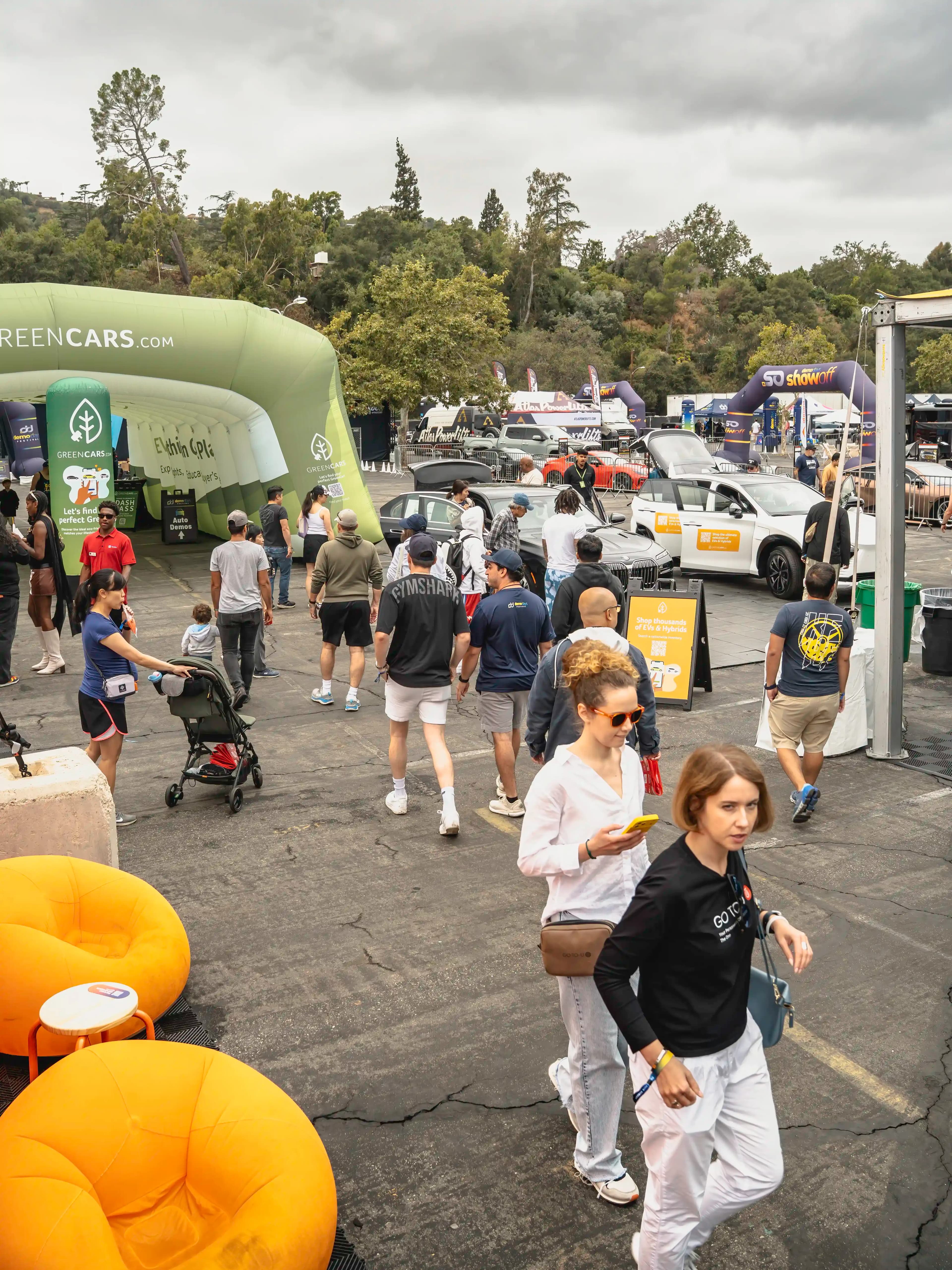 Una multitud pasa junto a carpas de GreenCars y vehículos eléctricos exhibidos en un área de demostración al aire libre.