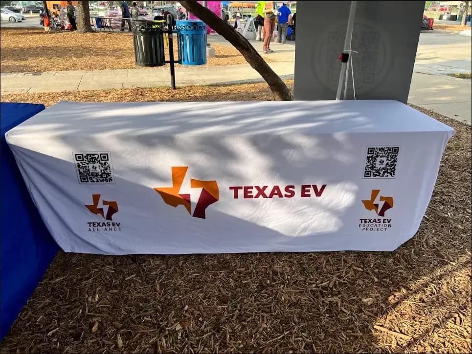 A table covered with a Texas EV Alliance banner at an outdoor event.