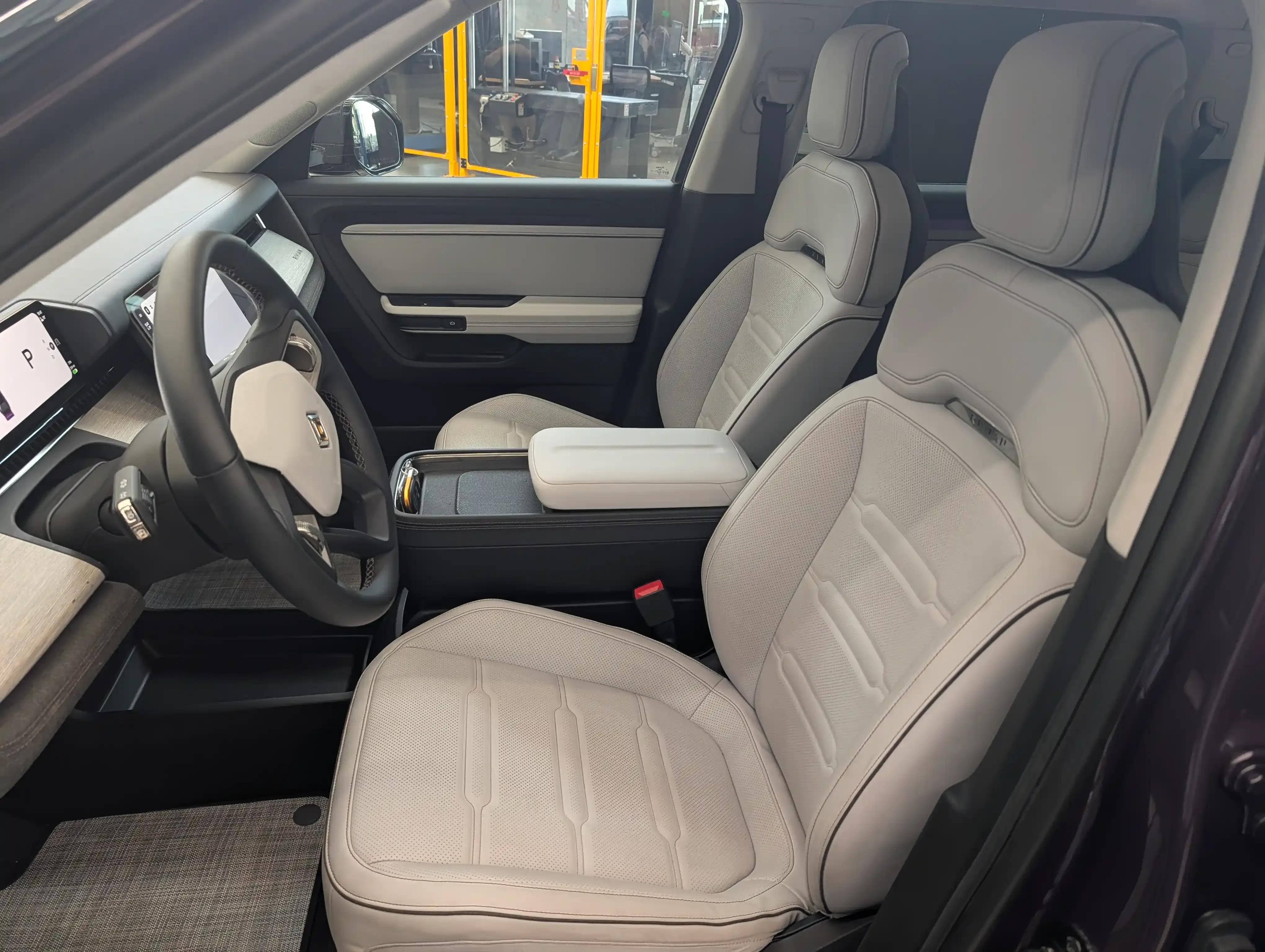 Front cabin of a Rivian R2 with a steering wheel, digital display, and light-colored seats.