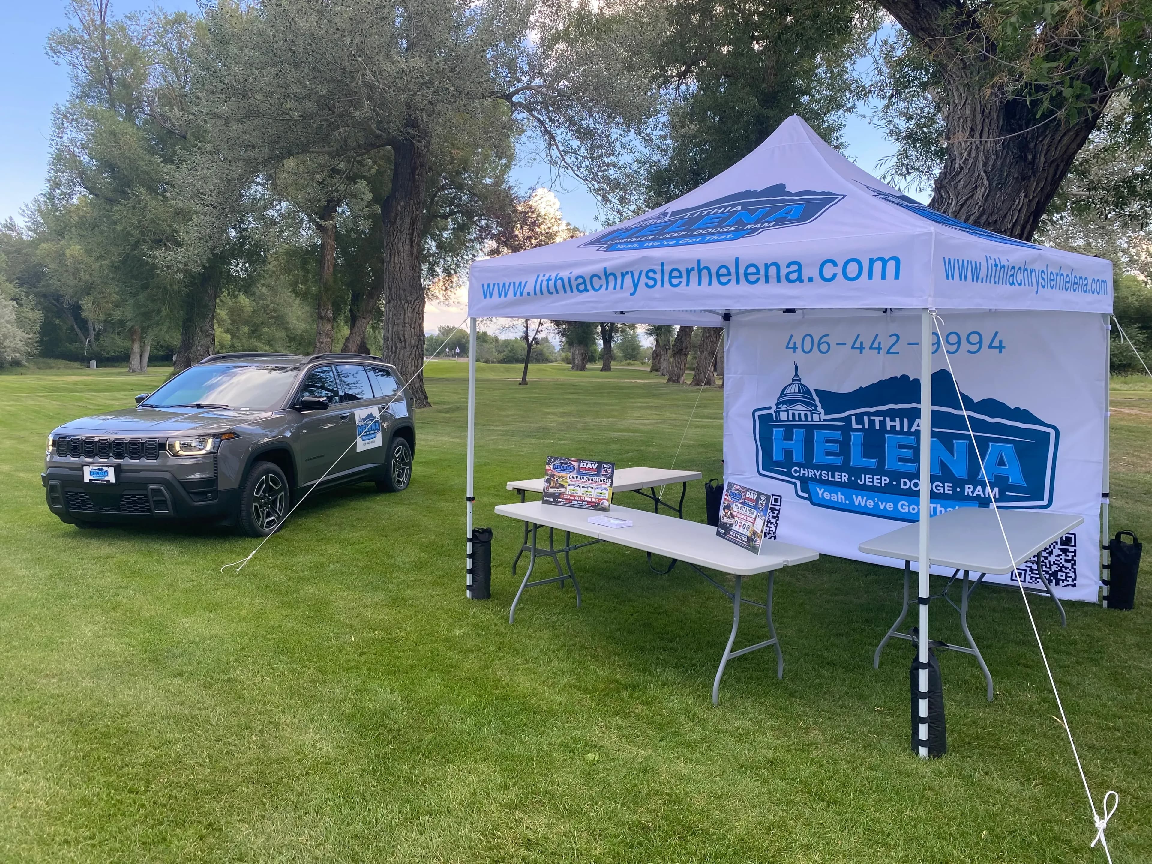 A gray Jeep Cherokee parked beside a Lithia Helena canopy tent on a grassy field.