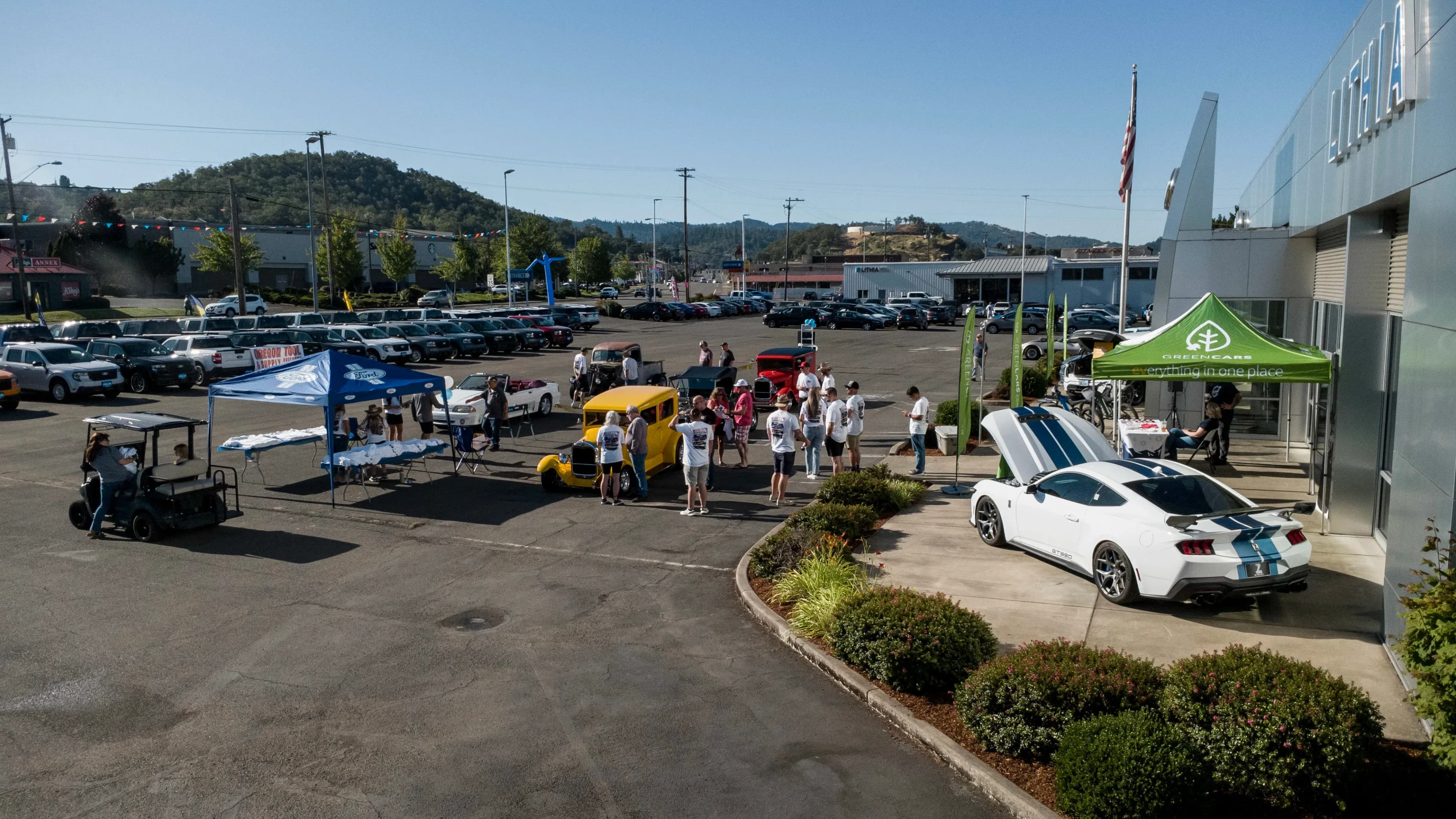 Vista amplia de un lote de exhibición de autos con carpas, personas y varios autos y camionetas clásicas.