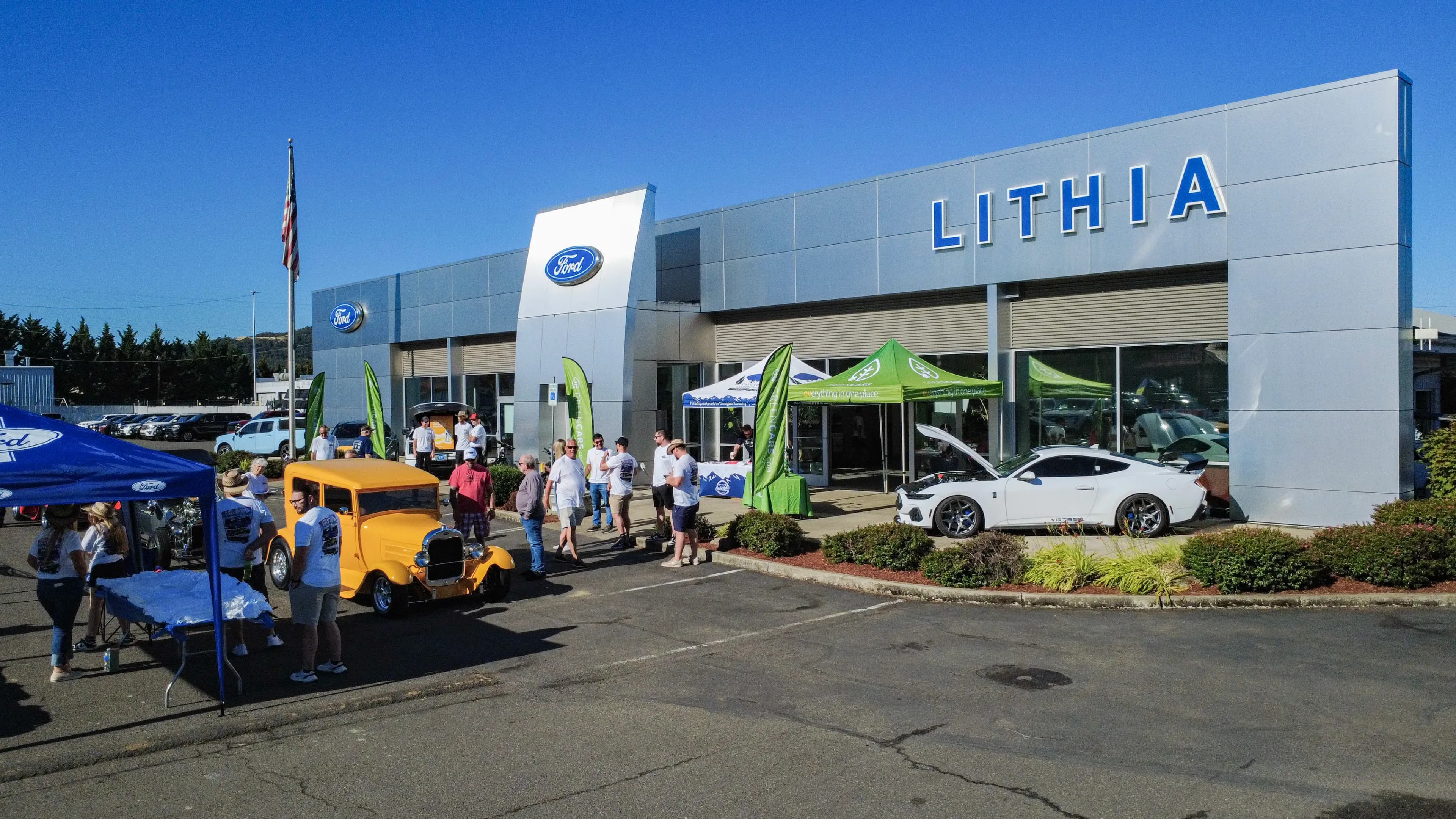 Vista exterior amplia de una concesionaria Lithia Ford con personas reunidas alrededor de dos autos estacionados y carpas del evento.
