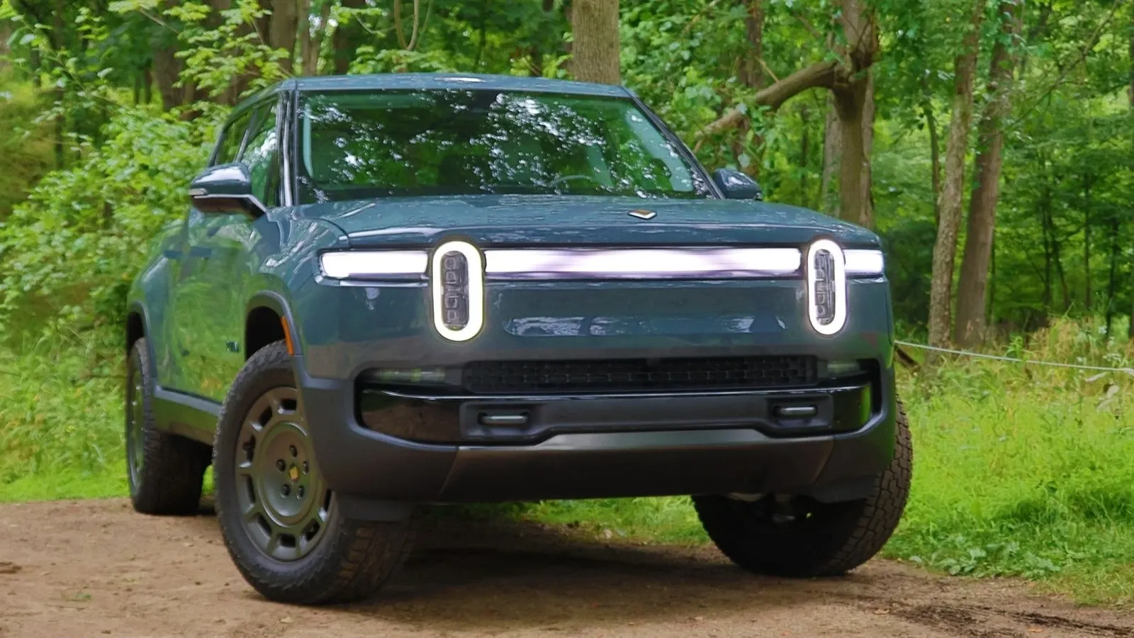 Blue 2026 Rivian R1T angled front image on a dirt road in front of a wooded area 