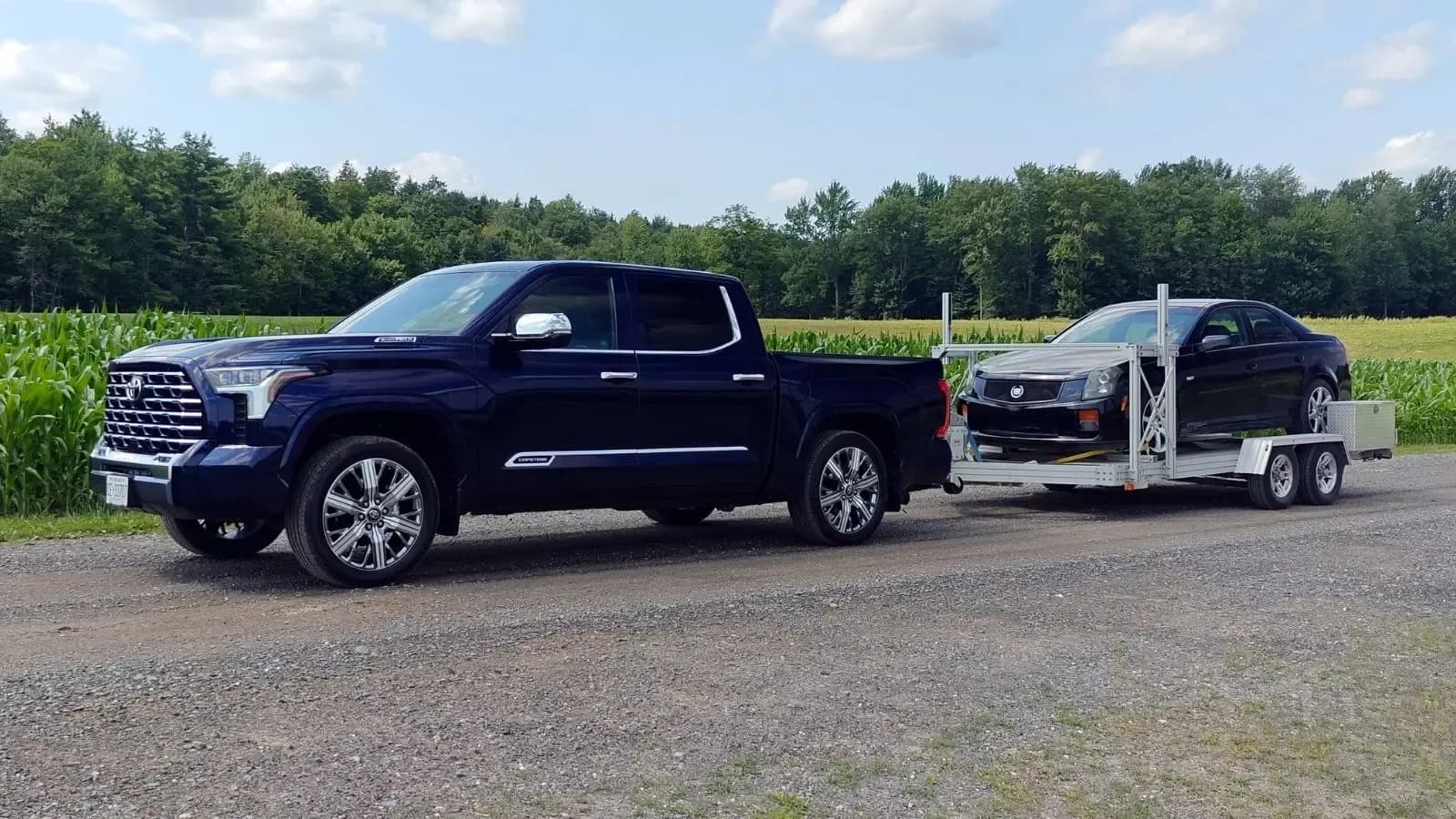 Blue 2026 Toyota Tundra Capstone Hybrid towing a black Cadillac car in front of a corn field