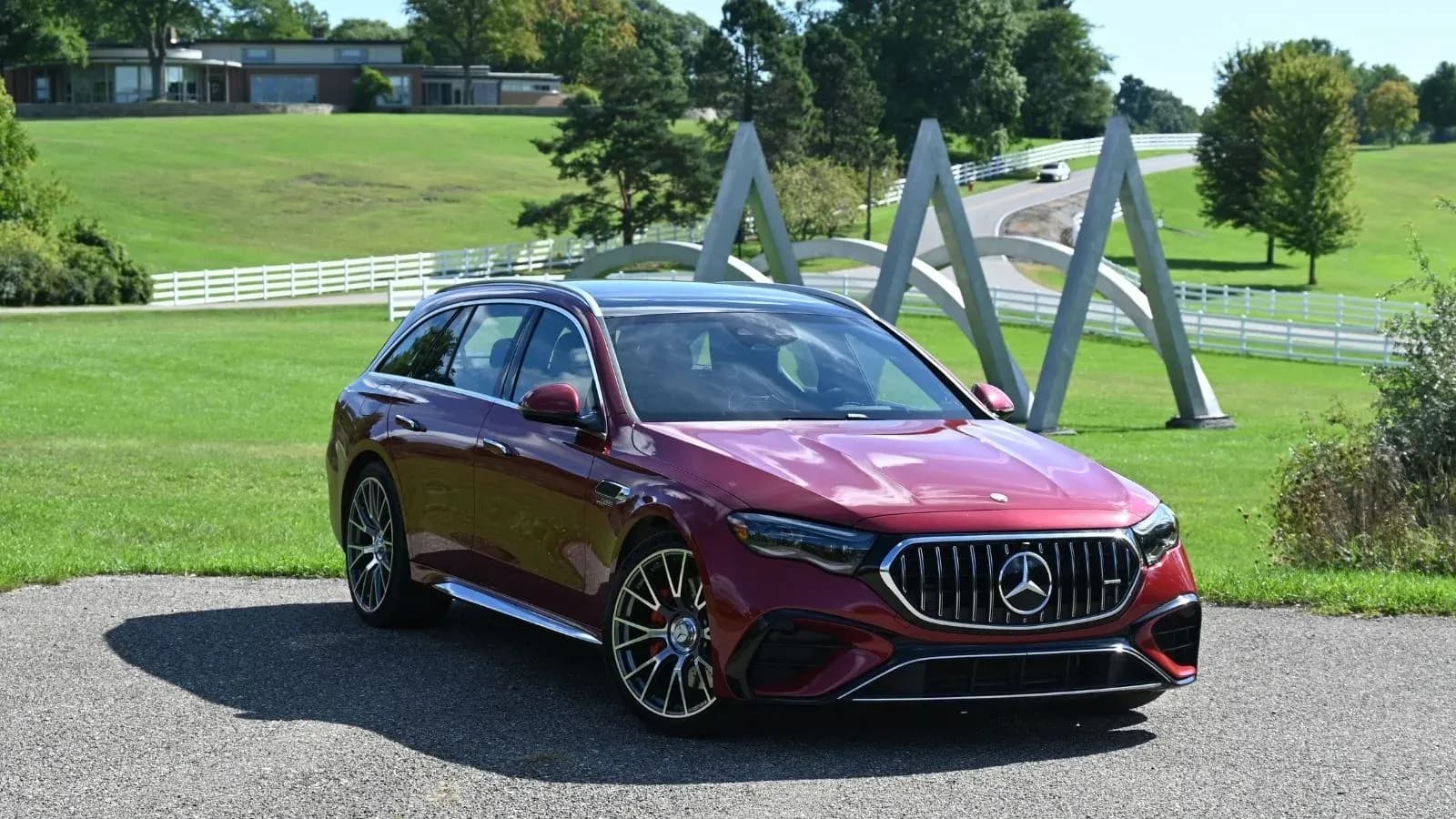 A red 2026 Mercedes-AMG E 53 Hybrid Wagon parked at angle in front of a sculpture and landscaped property